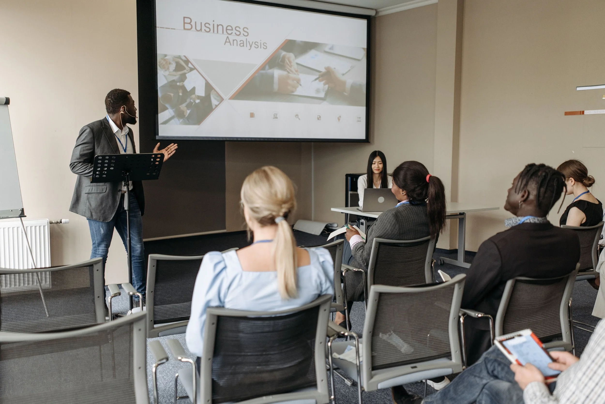 A man in a suit presenting a slide titled 'Business Analysis' to a group of diverse attendees in a conference room.