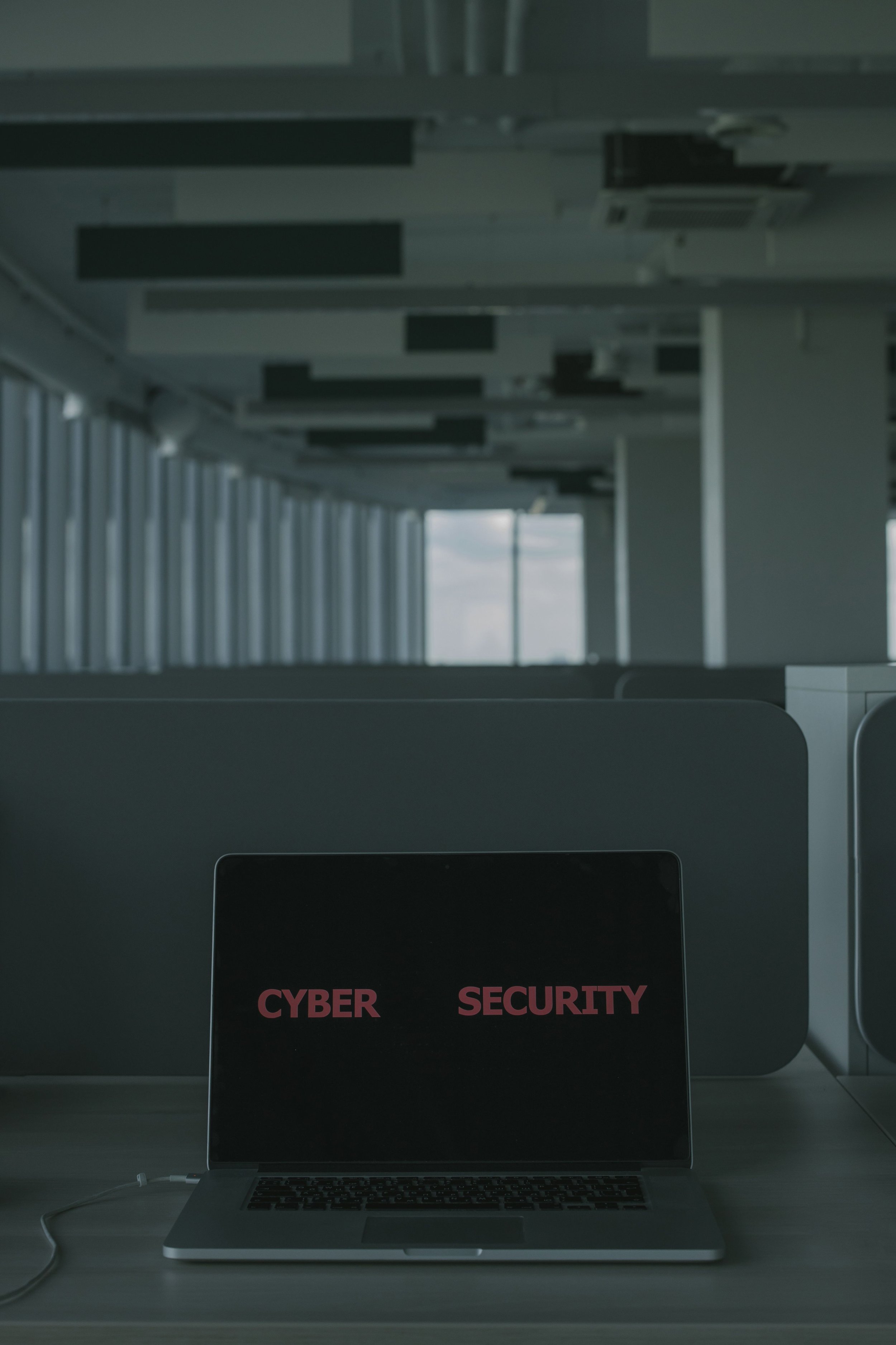 Laptop on a desk with a screen displaying the words 'CYBER SECURITY' in pink letters, in an office setting with large windows and ceiling air conditioning units.