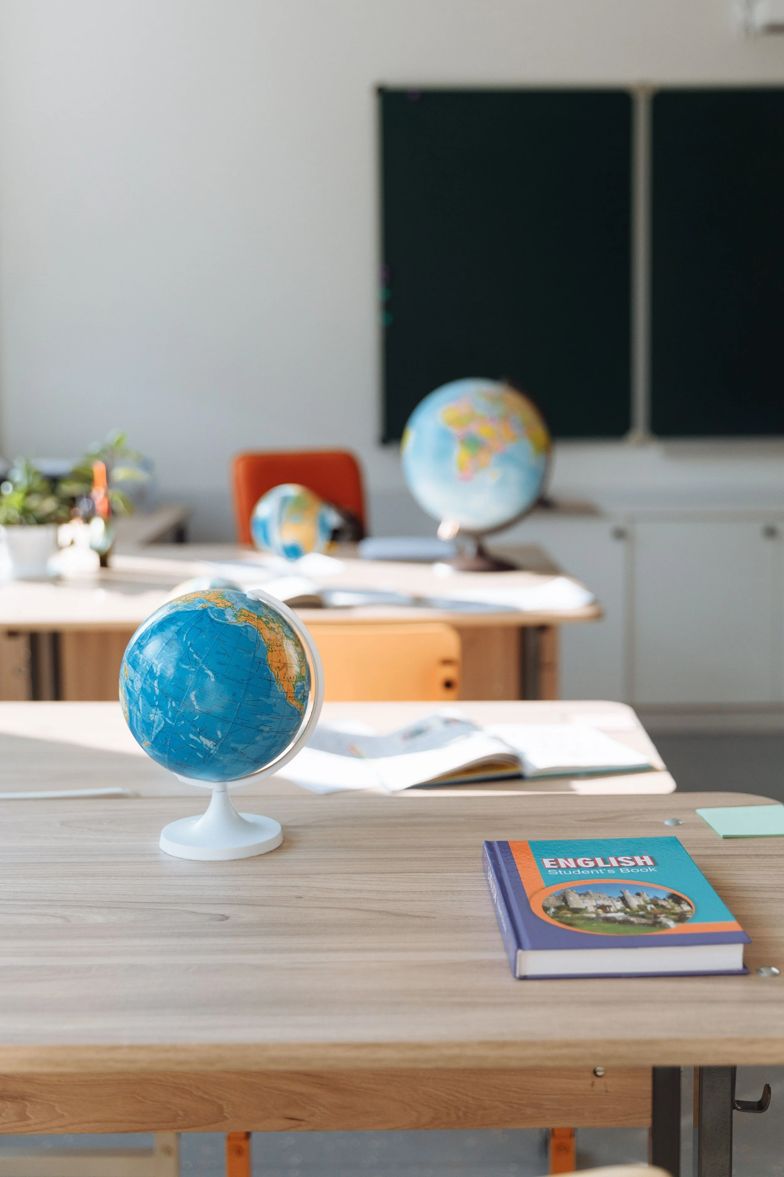 A classroom desk with a small globe, an English student’s book, and an open notebook, with more globes and a blackboard in the background.