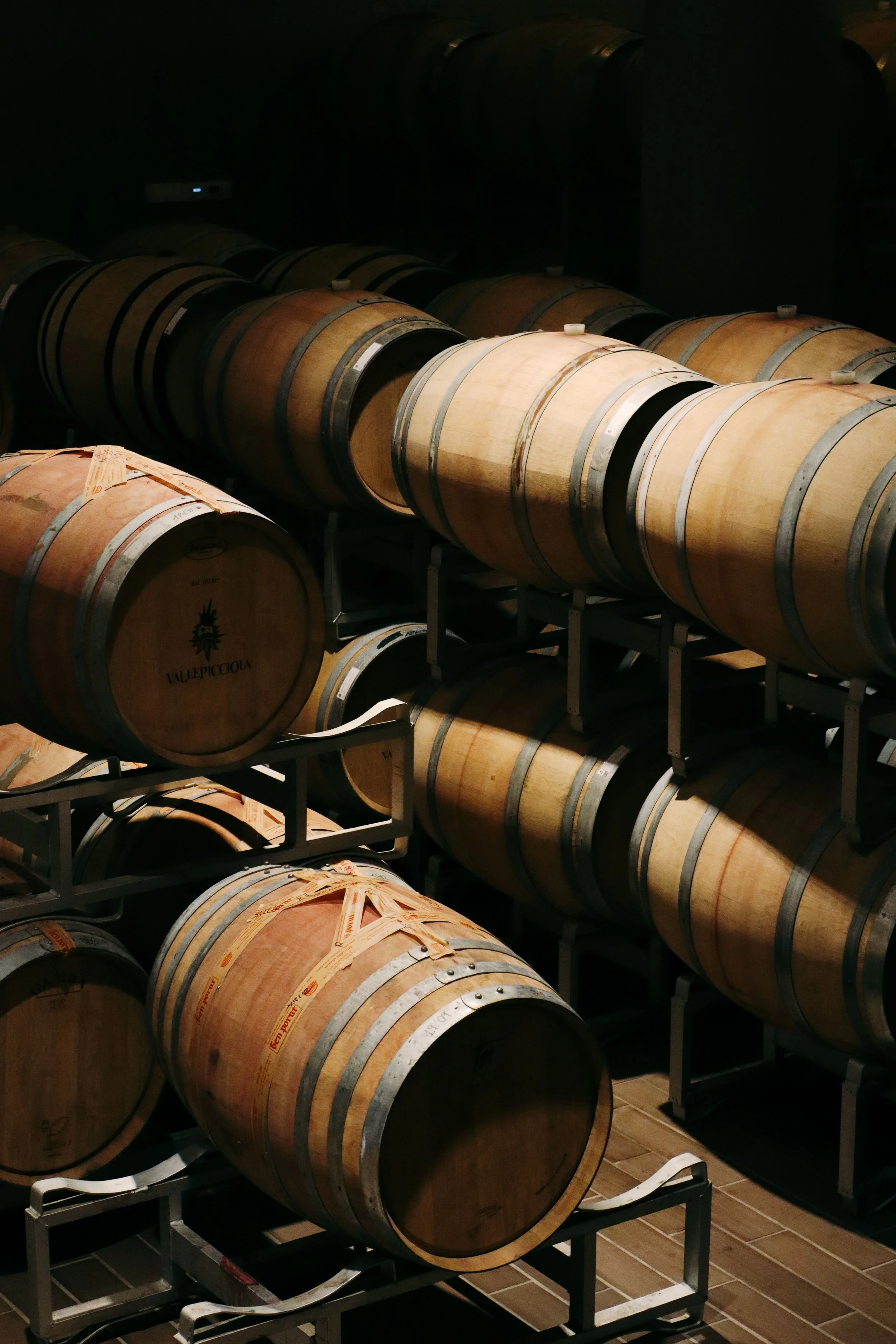 A wine cellar with multiple wooden wine barrels stored on metal racks in a dimly lit room.
