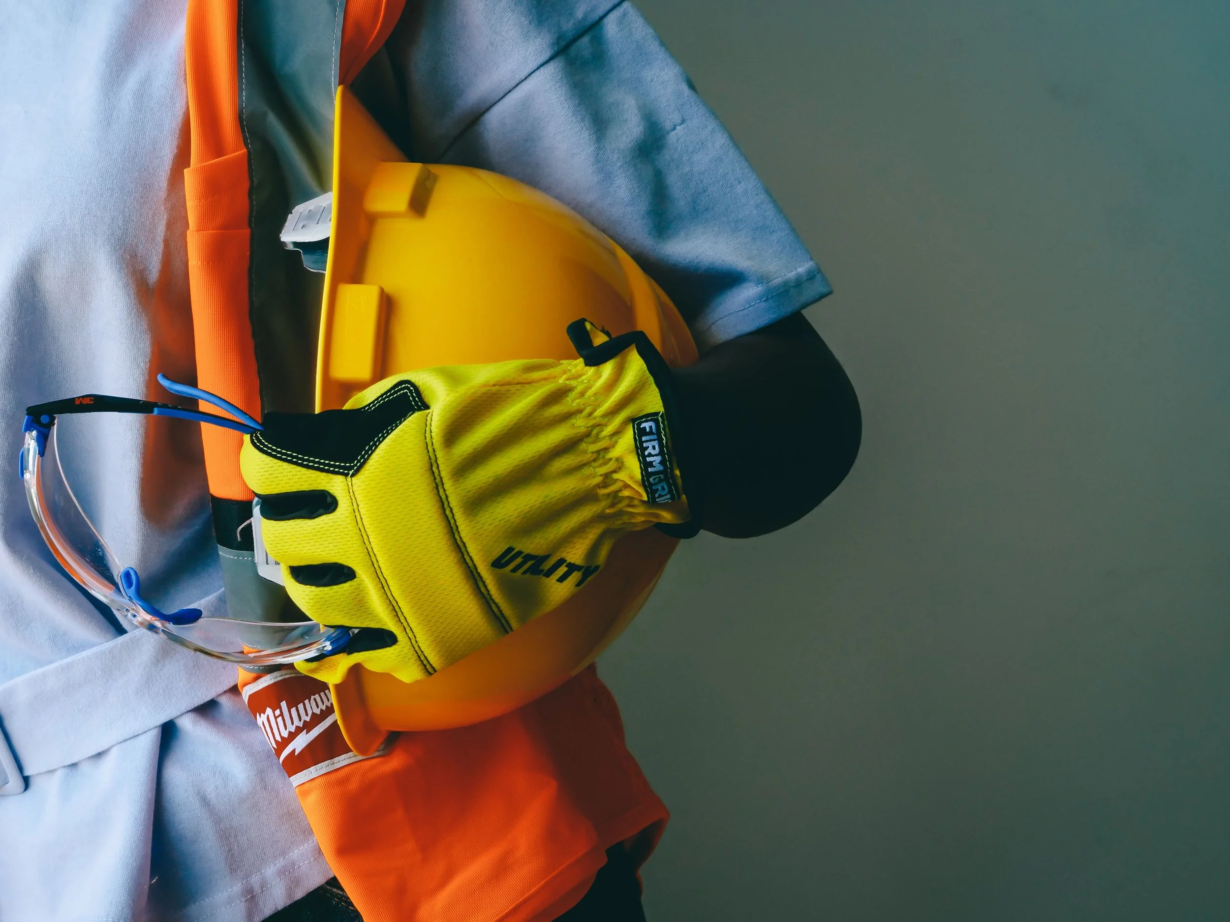 A close-up of safety gear including yellow work gloves, a yellow hard hat, and part of a gray jacket with an orange shirt underneath.