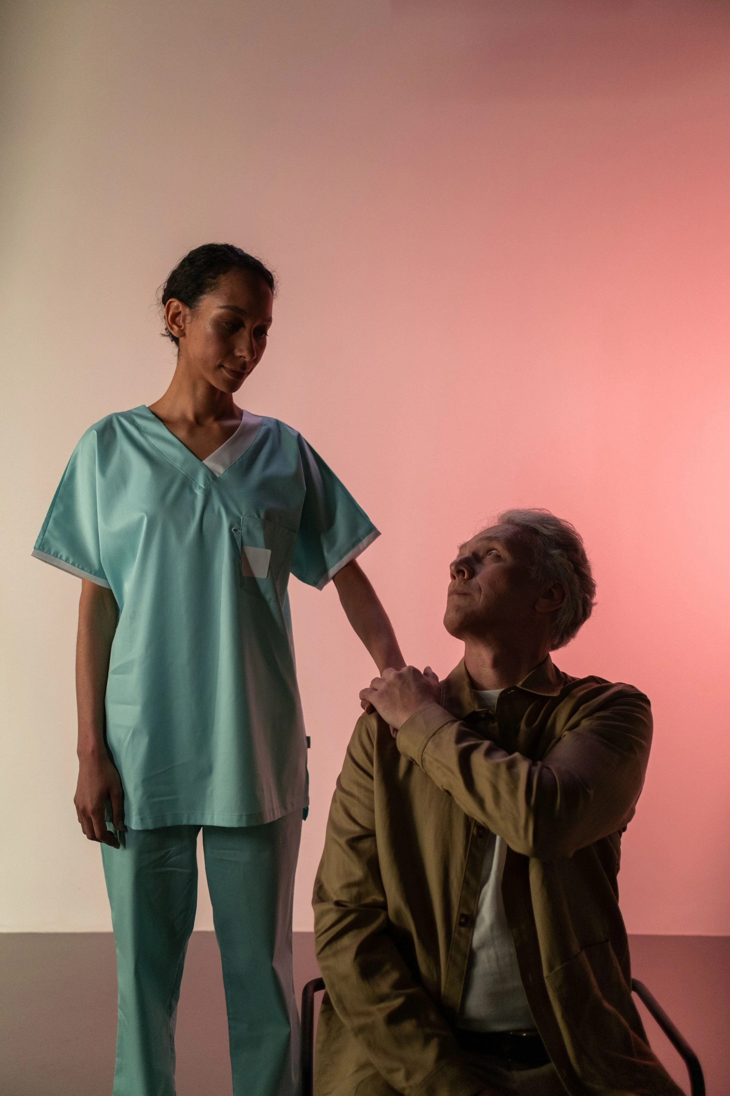 A middle-aged woman in a nurse's uniform standing next to an elderly man with gray hair sitting in a chair. The woman is touching the man's shoulder and they are looking at each other in a scene with soft pink and purple lighting.