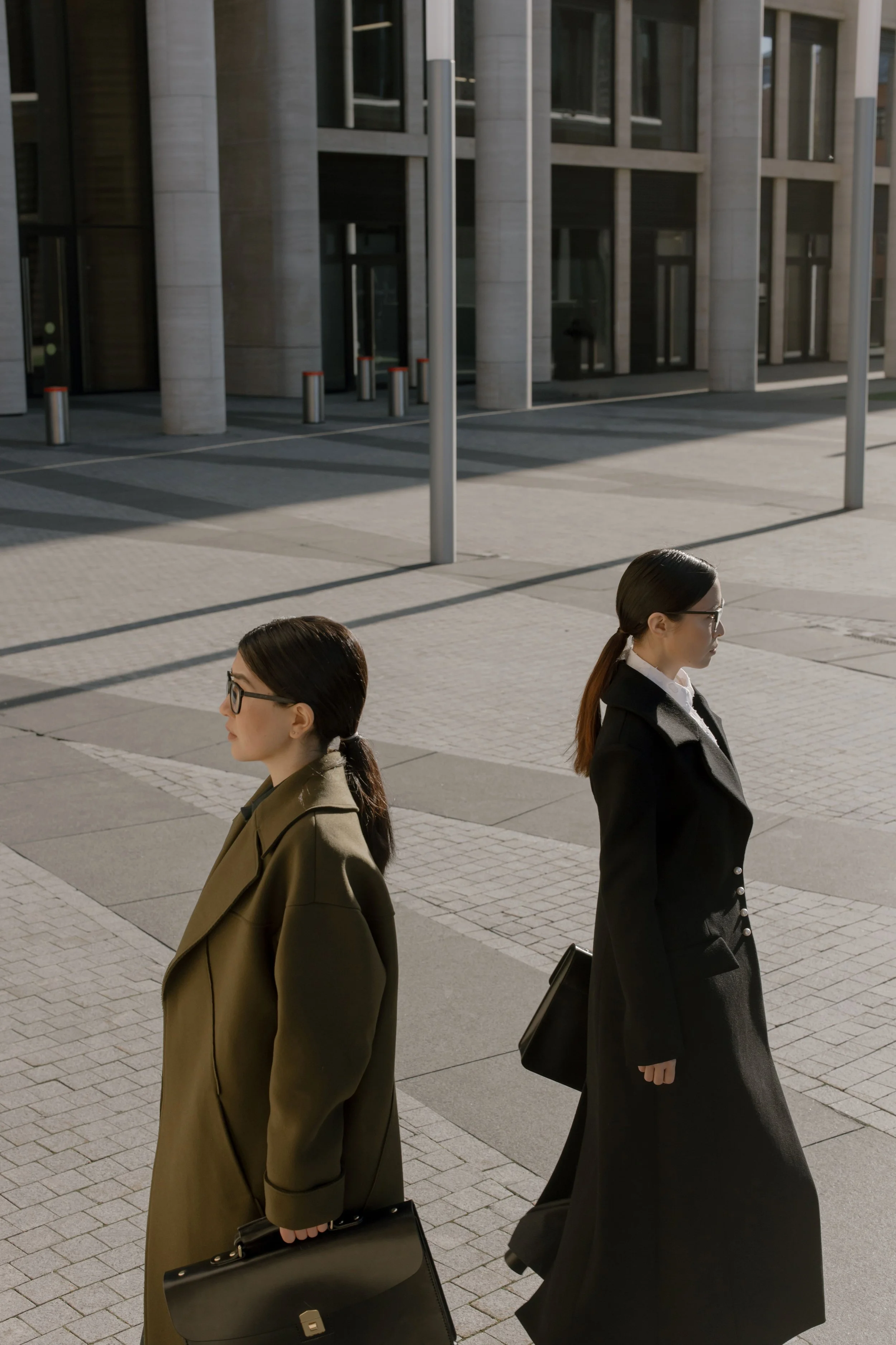 Two women walking outside near a modern building with large glass windows. One woman is wearing a beige coat and glasses, holding a black briefcase. The other woman is dressed in a black coat with a matching umbrella, and also wears glasses.