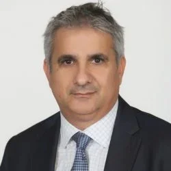 Professional headshot of a middle-aged man with gray hair, wearing a dark suit, white shirt, and patterned tie, against a plain light background.