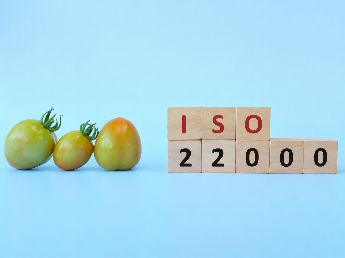 Three heirloom tomatoes of different sizes and colors next to wooden blocks spelling 'ISO 22000' against a light blue background.