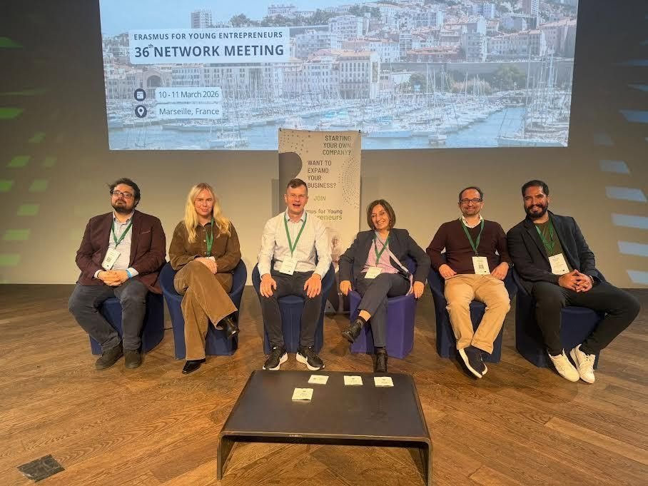 Six people sitting on chairs on a stage at a conference, with a large screen behind them displaying information about the Erasmus for Young Entrepreneurs 36th Network Meeting in Marseille, France, dated March 10-11, 2026.
