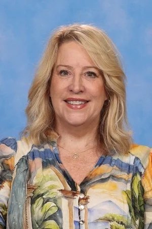Portrait of a smiling woman with blonde hair wearing a colorful, patterned blouse against a blue background.
