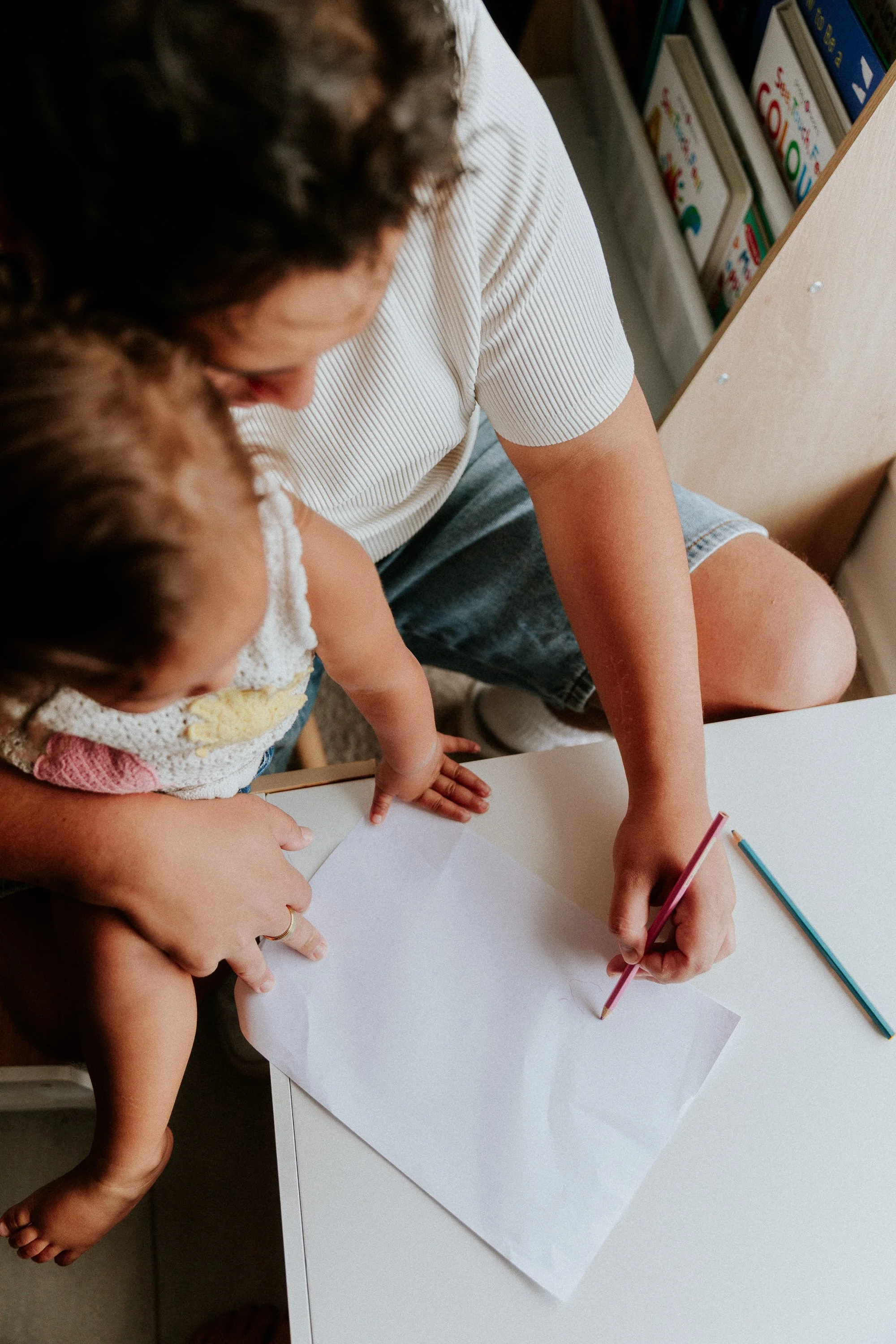 A woman and a child are sitting at a white table, drawing or writing on a blank sheet of paper with colored pencils. The woman is wearing a white and black striped shirt, and the child is wearing a colorful crocheted top. Books are visible on a shelf in the background.