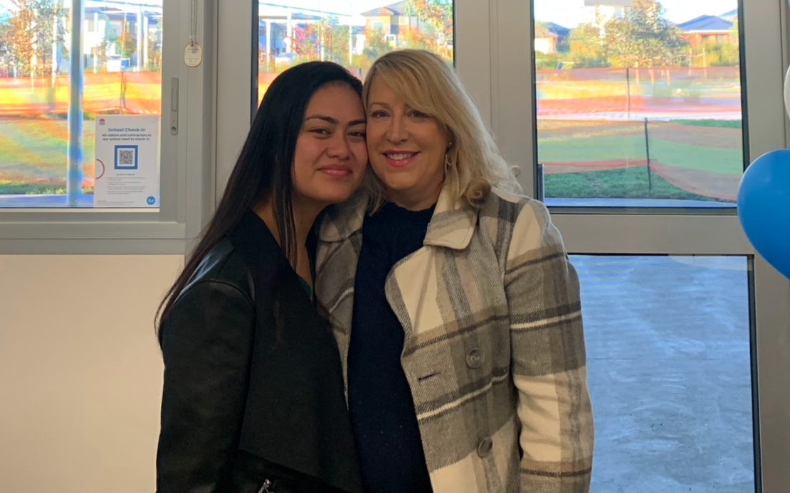 Two women standing close together, smiling, with large windows behind them showing an outdoor sports field with trees and houses.