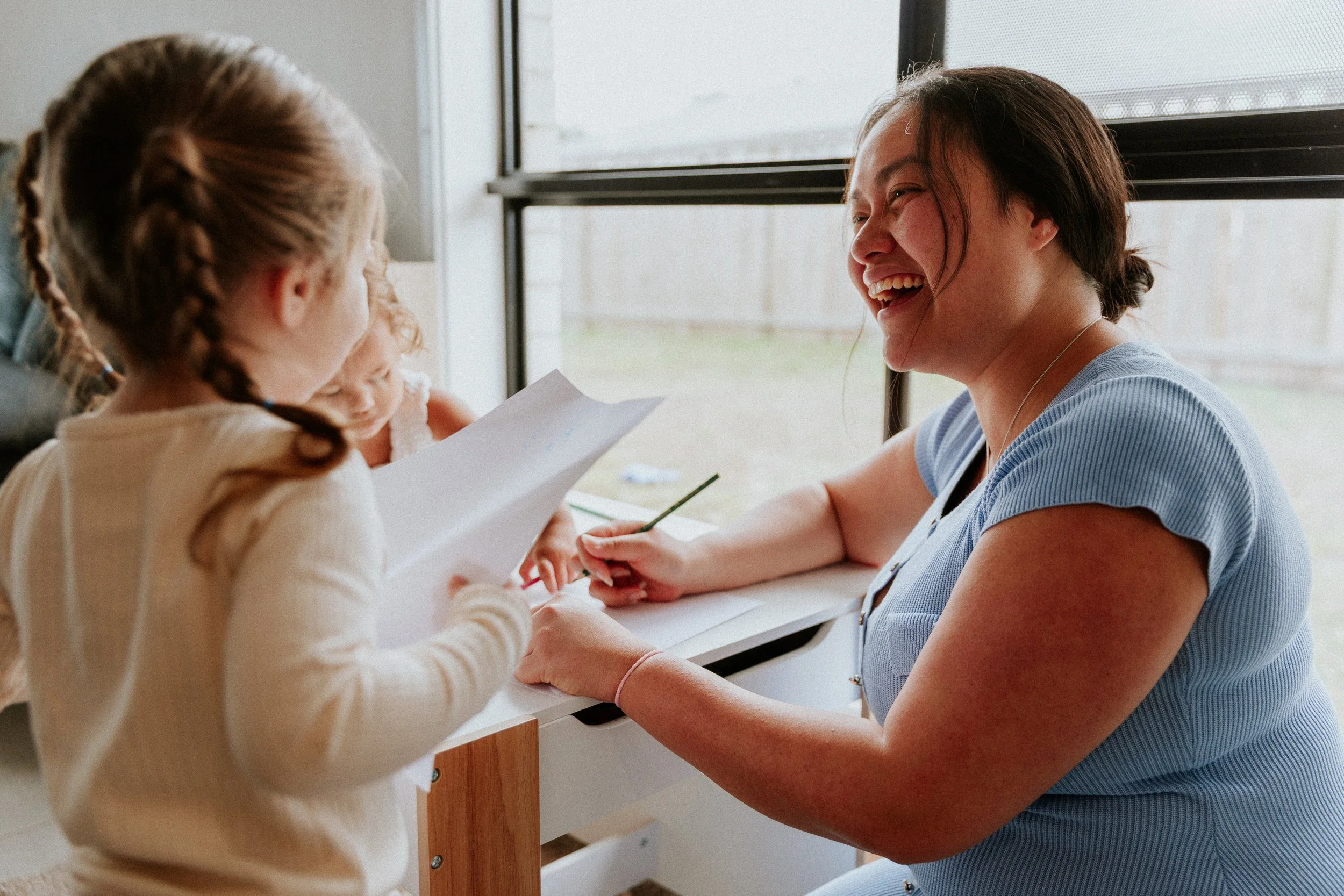 A woman and two young girls smiling and engaging in an activity together at a table near a window.
