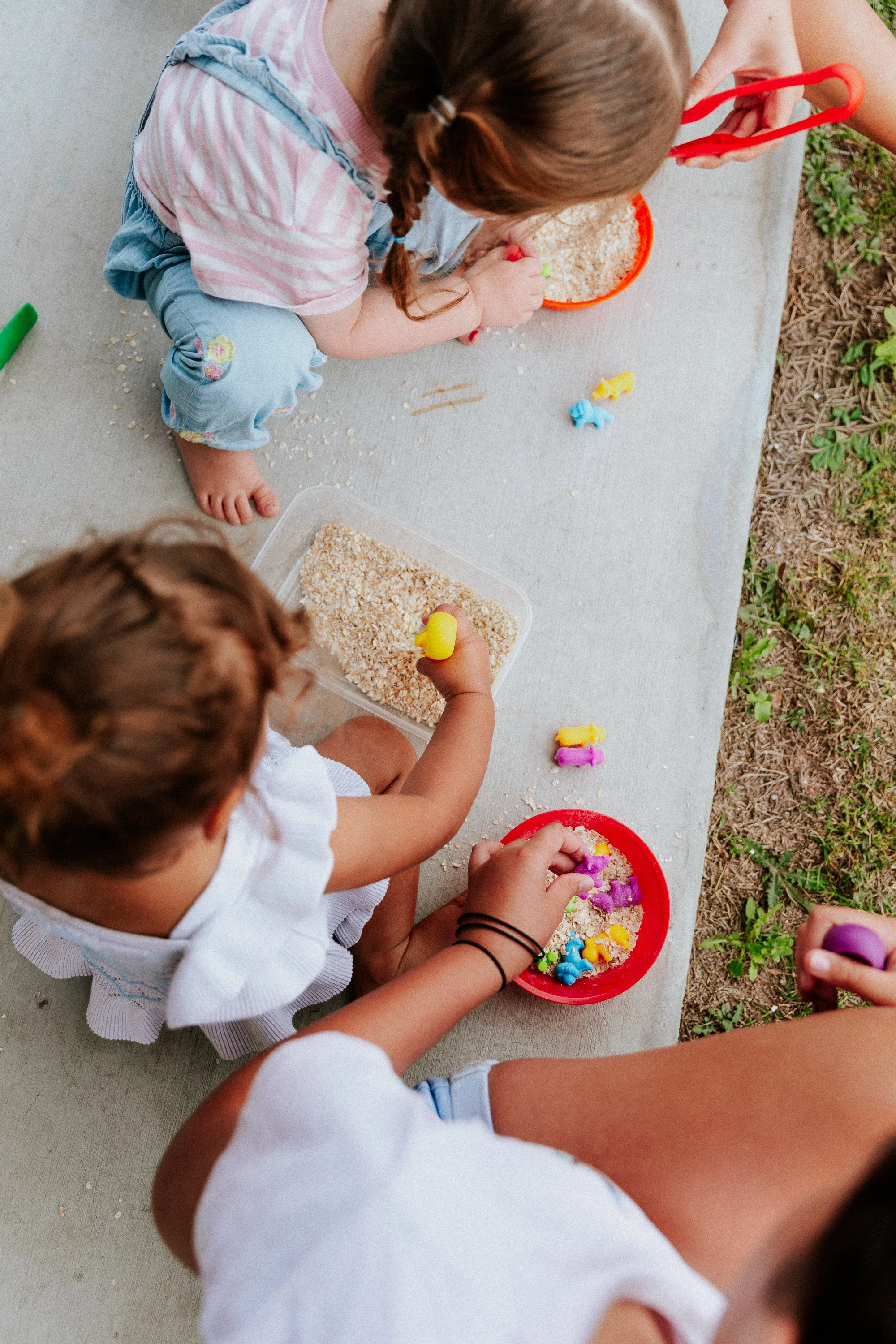 Children sitting on a bench outside, playing with colorful toys and sand.