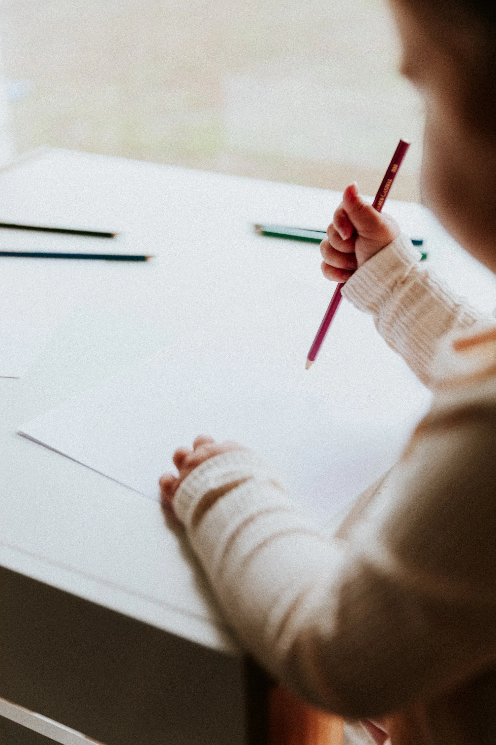 A young girl wearing a beige sweater holding a pink colored pencil over a white sheet of paper.