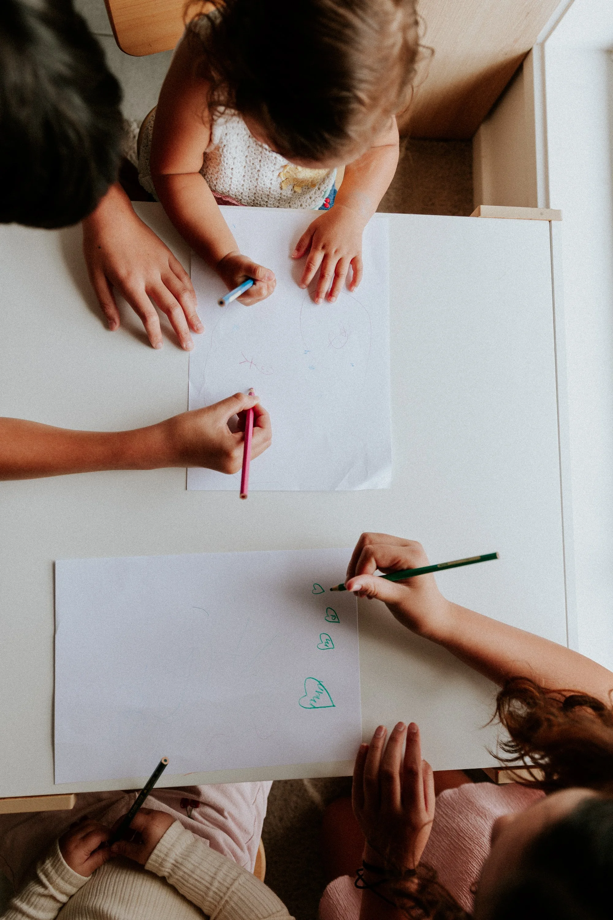 Four children drawing and writing on white sheets of paper at a table, with colored pencils in their hands.