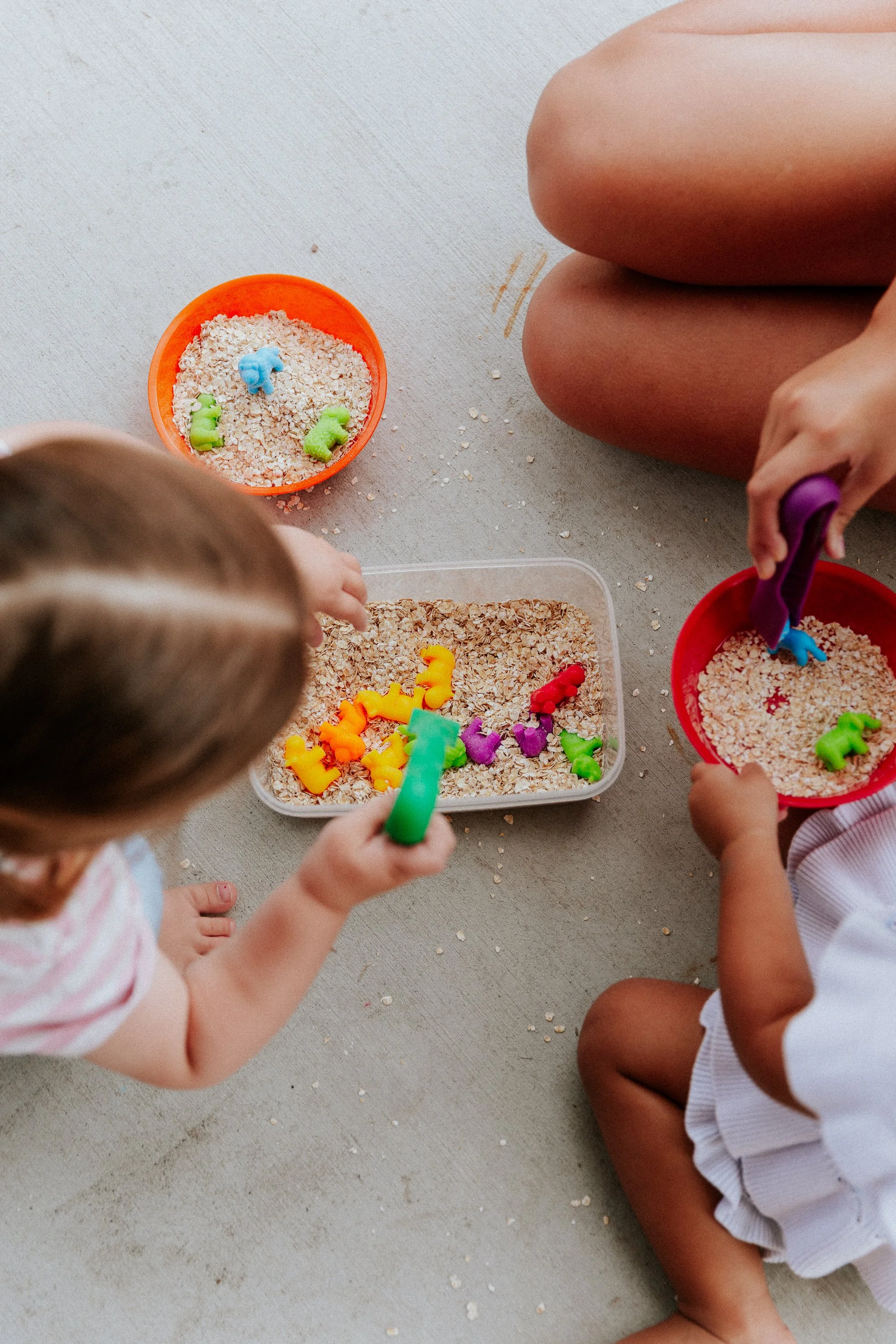 Children playing with colorful animal-shaped toys in a container of oats, sitting on a beige surface.