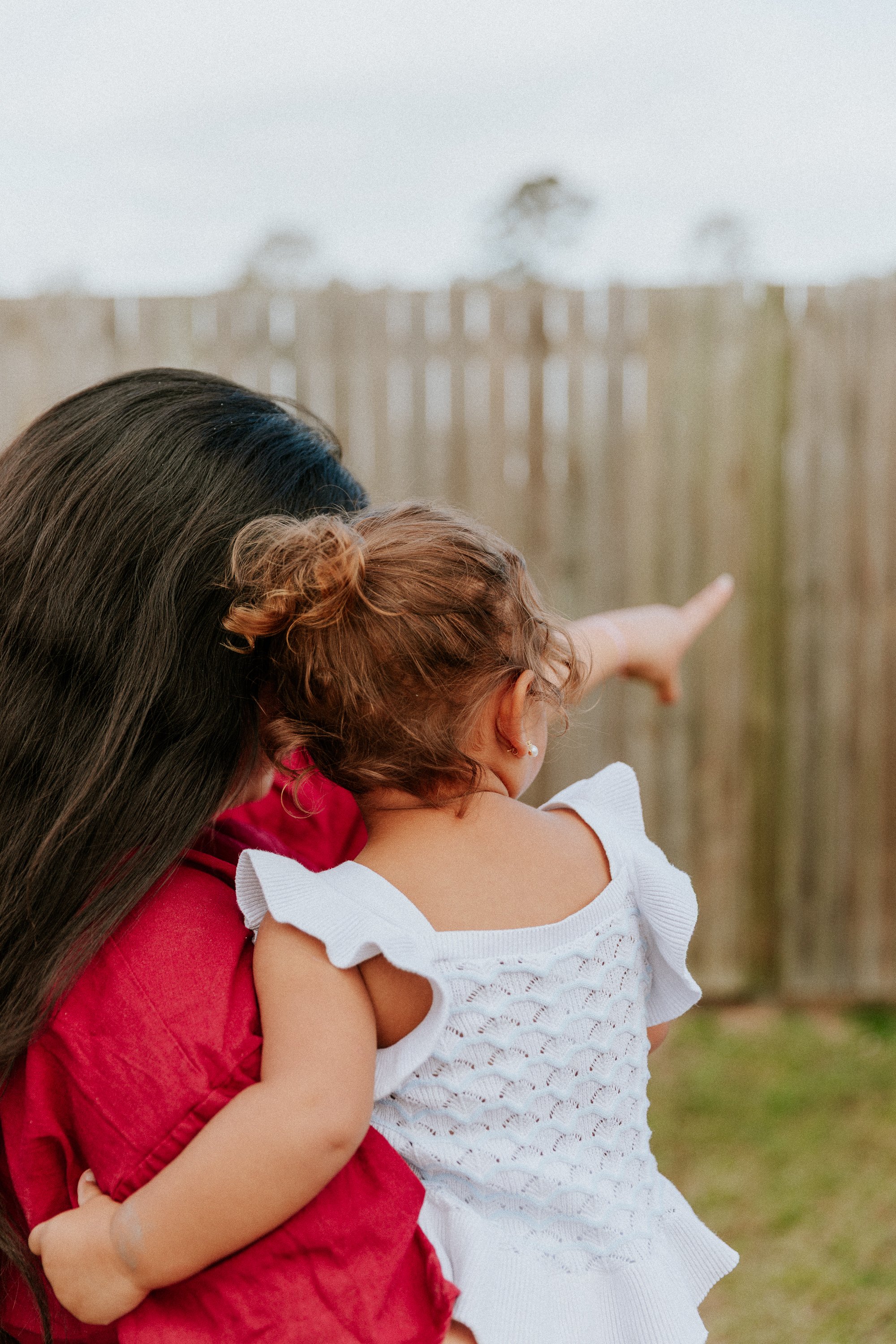 A woman with long dark hair and a child with curly red hair, both pointing at something outside, in a backyard with a wooden fence and trees in the background.