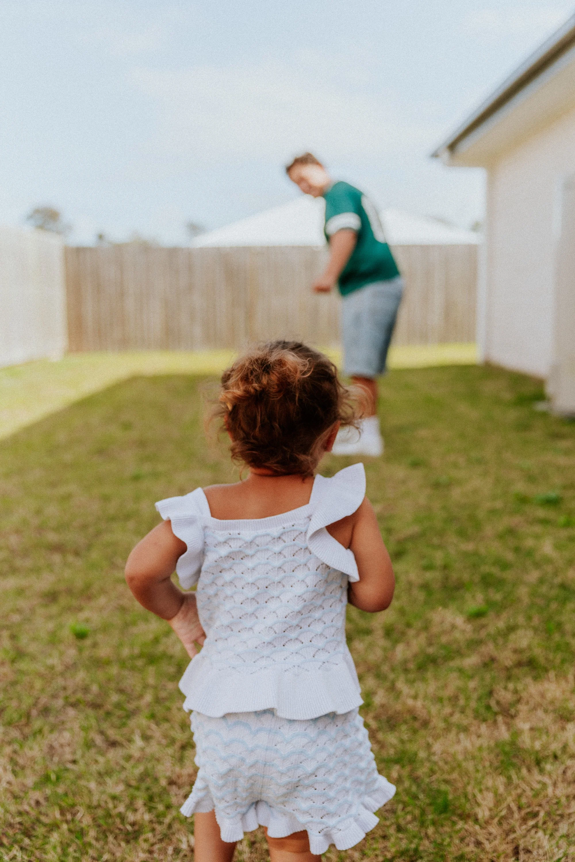 Young girl in white ruffled dress standing in a backyard, looking at a boy in a green shirt and gray shorts playing nearby.