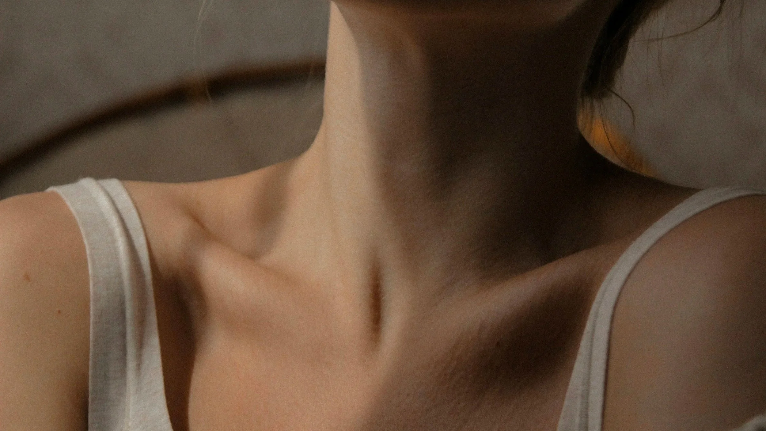Close-up of woman's neck, shoulder, and a part of her upper chest. She is wearing a white tank top, and the background appears to be neutral.