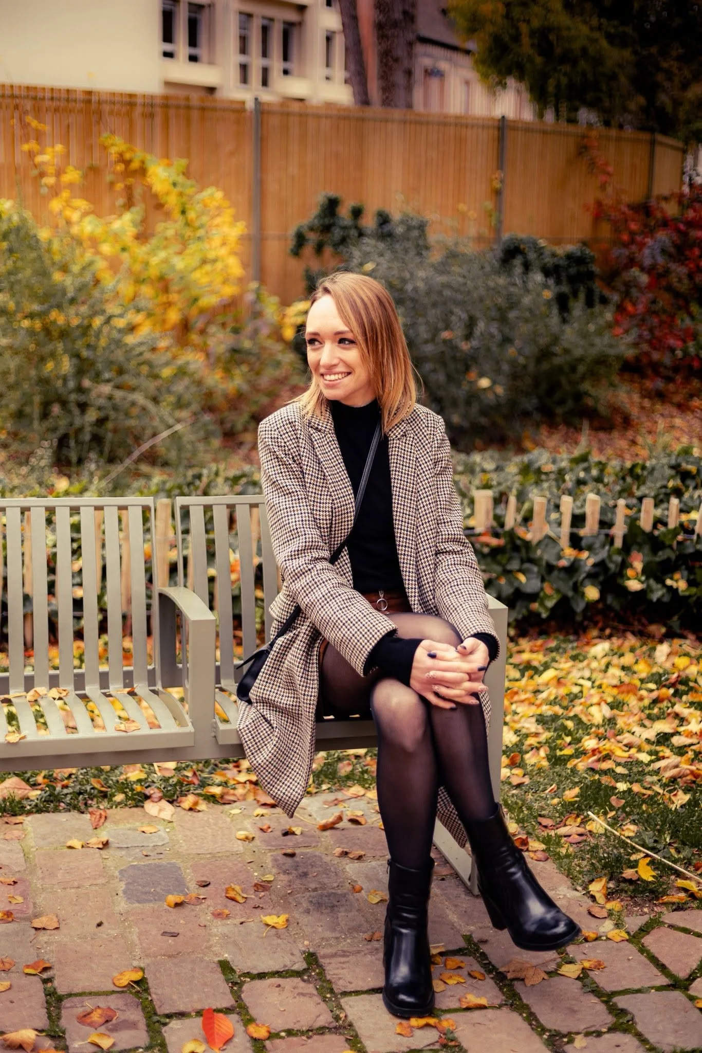 Une femme assise sur un banc en bois dans un parc automnal, portant un manteau à carreaux, une robe noire, des collants en nylon et des bottes en cuir, souriant et regardant vers la droite, entourée de feuilles mortes.