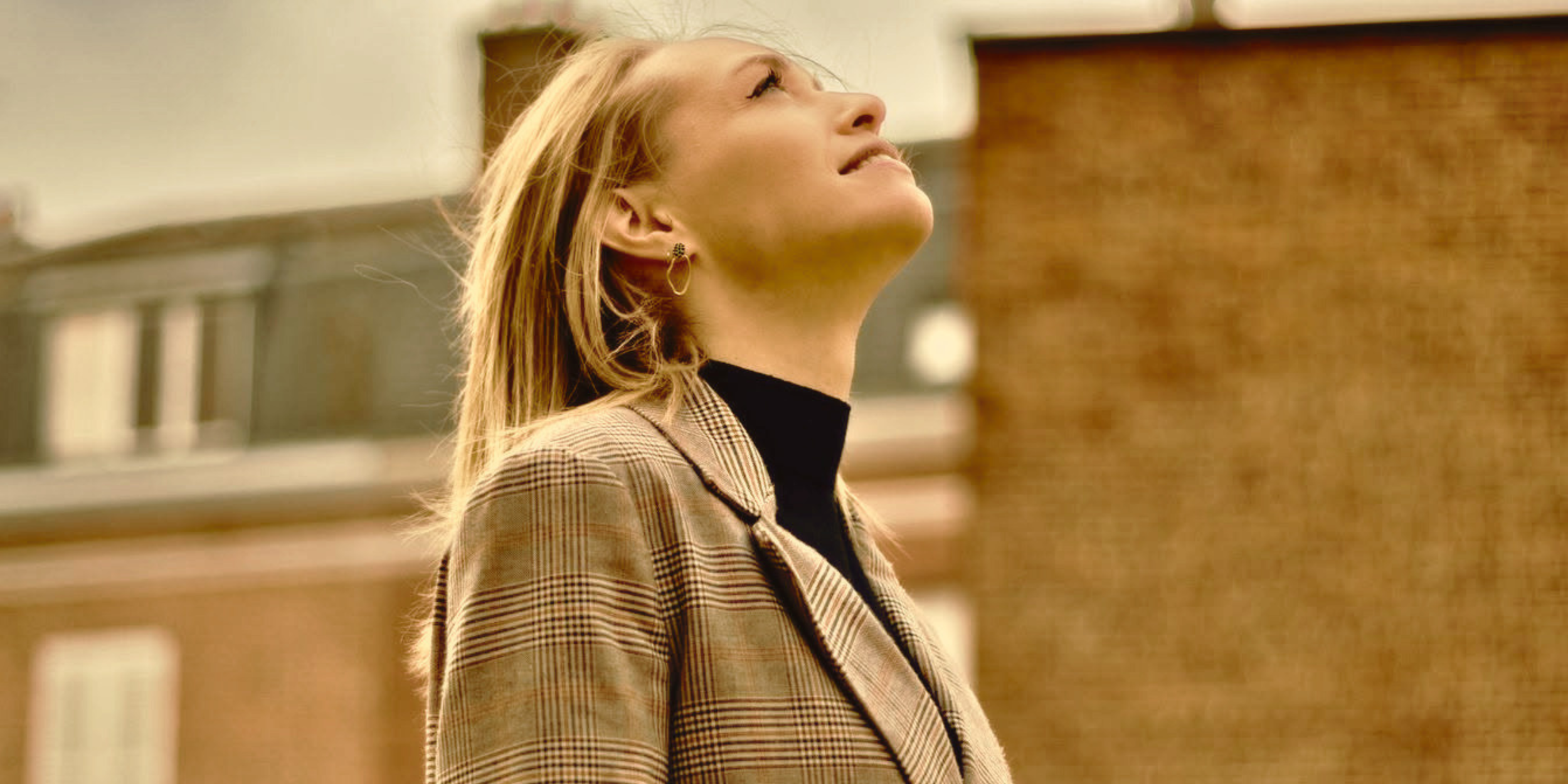 Une femme blonde regarde vers le haut, portant une veste à carreaux et un t-shirt noir, dans un environnement urbain avec des bâtiments en arrière-plan.