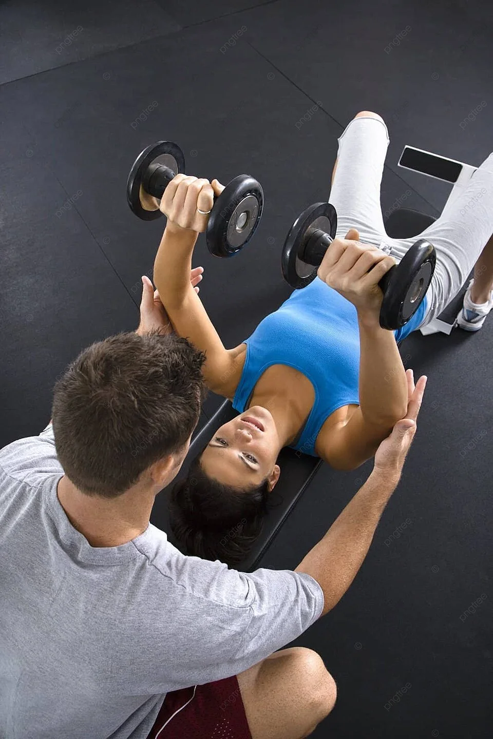 A woman lying on a gym bench lifting dumbbells while a trainer assists her.