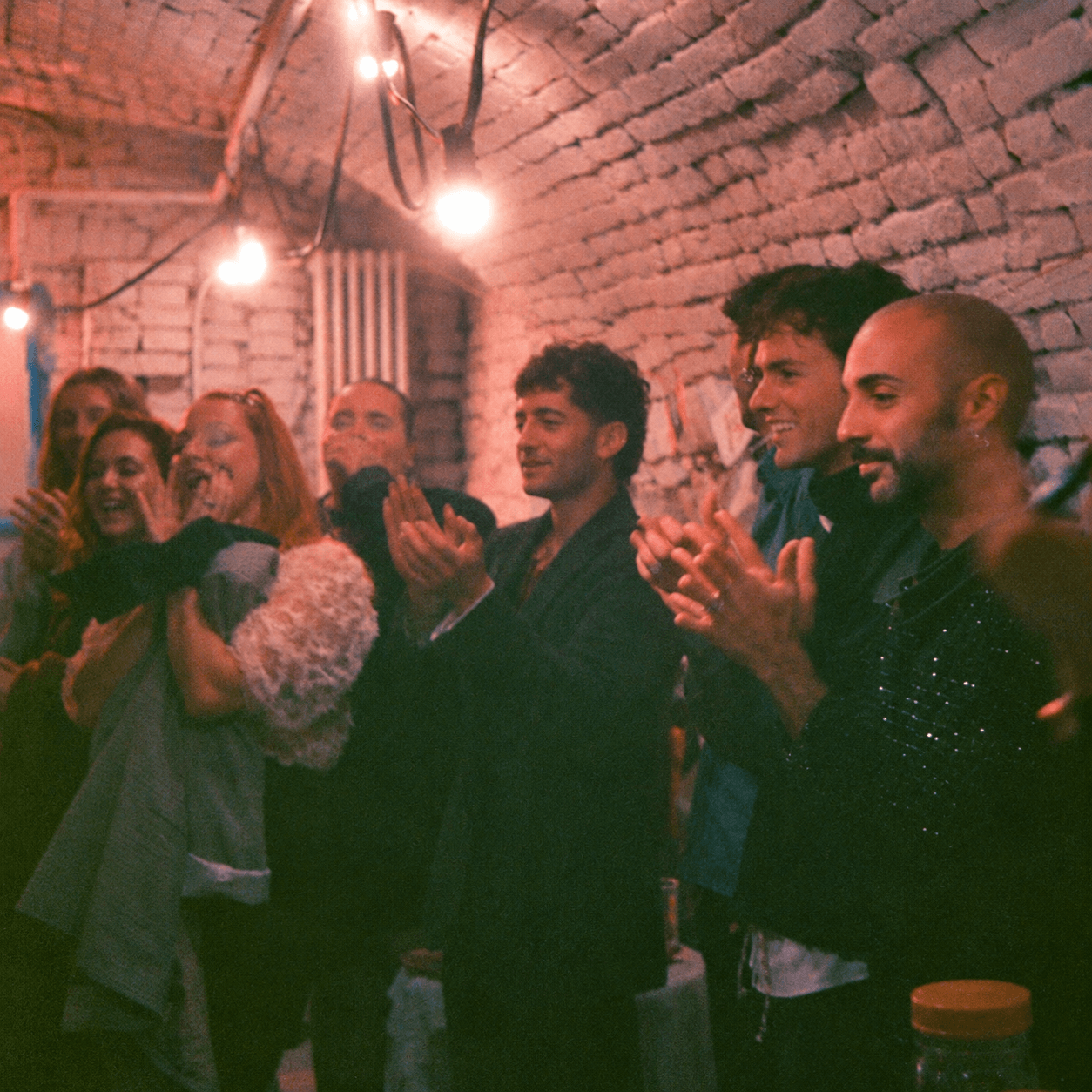 Un gruppo di persone sorride e applaude in un ambiente con pareti di mattoni e luci calde. Casa delle Stelle, ambiente e associazione queer a Brescia