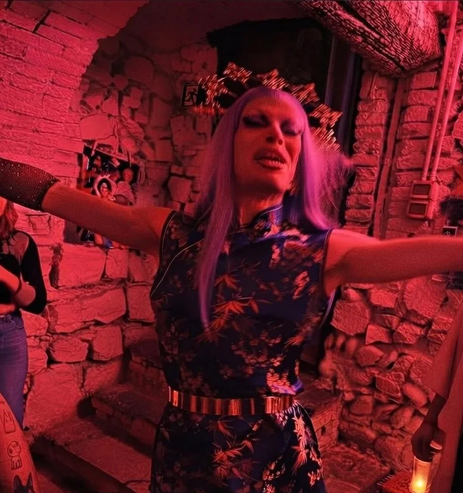 Drag queen con abito floreale in un locale con pareti di pietra rossa, luci soffuse e decorazioni da party. Casa delle Stelle, ambiente e associazione queer a Brescia