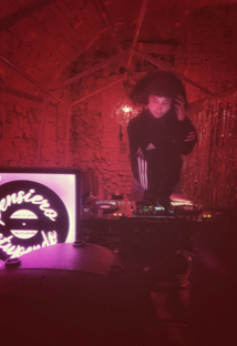 DJ Fgiaca che suona in un locale Casa delle Stelle, ambiente e associazione queer a Brescia, con decorazioni rosse e un cartellone luminoso con il testo Pensiero stupendo