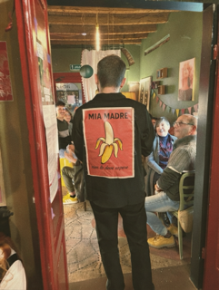 Persona con giacca che ha sulla schiena una stampa di una banana e la scritta 'Mia Madre' in un locale affollato. Casa delle Stelle, ambiente e associazione queer a Brescia