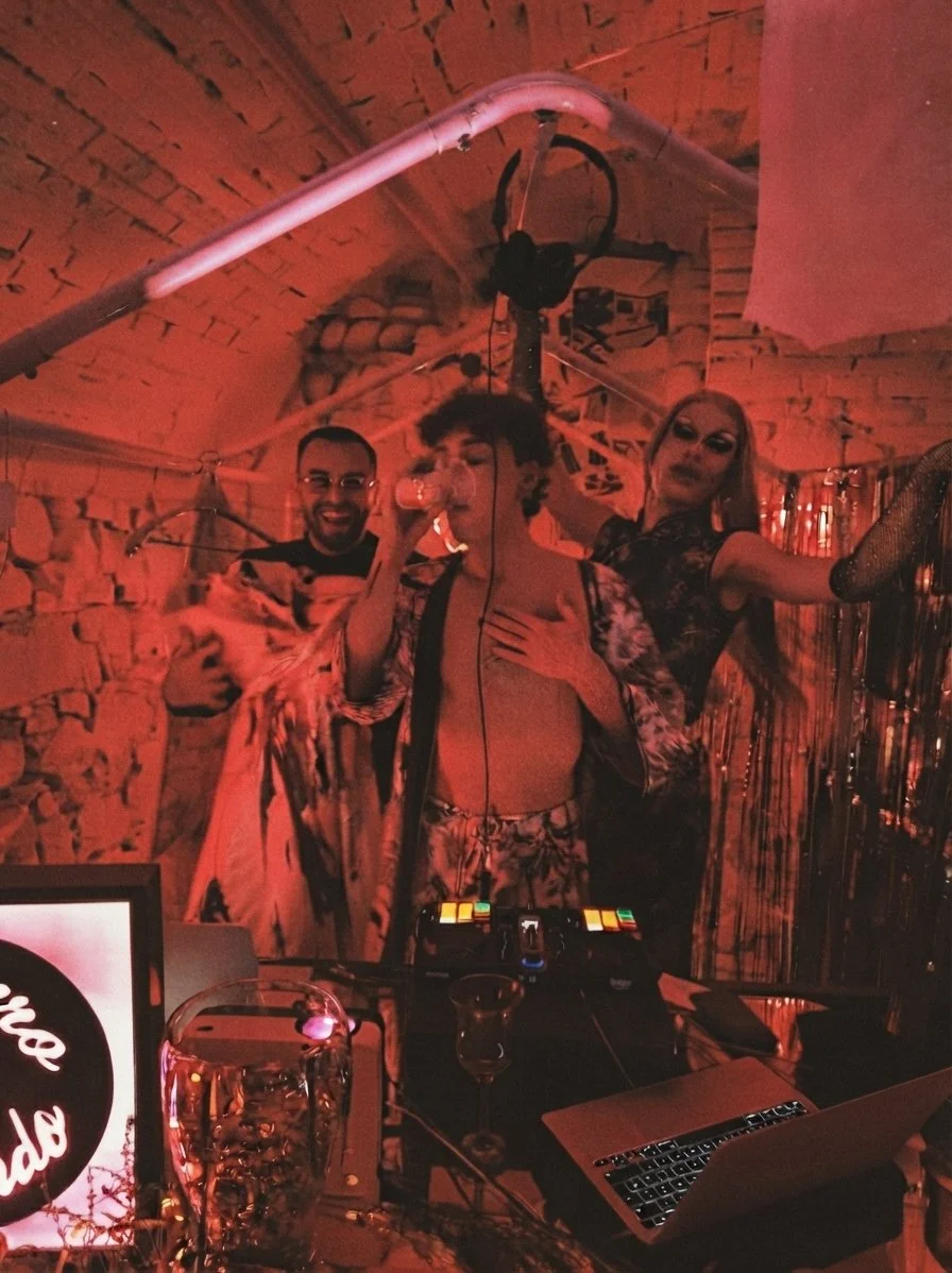 Tre persone dj set in un ambiente con luci rosse. Sul tavolo davanti ci sono un laptop, due bicchieri e un bicchiere con una bevanda.