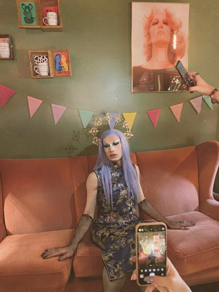 Drag Queen con trucco drammatico, capelli viola e abiti decorativi, seduta su un divano in una stanza decorata con bandierine colorate, una parete verde con quadri e un grande poster di una donna con capelli arancioni. Casa delle Stelle a Brescia