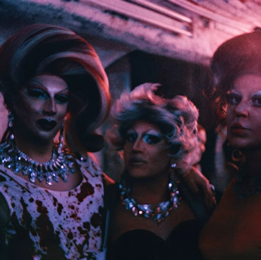 Tre Drag Queen con trucco audace e abiti eleganti in un contesto con luci soffuse. Casa delle Stelle, ambiente e associazione queer a Brescia. Salamandra Sisters