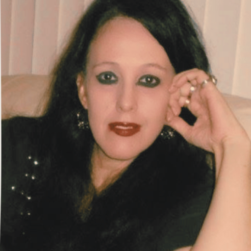 A woman with long dark hair, wearing makeup and a black top, is sitting indoors and resting her head on her hand.