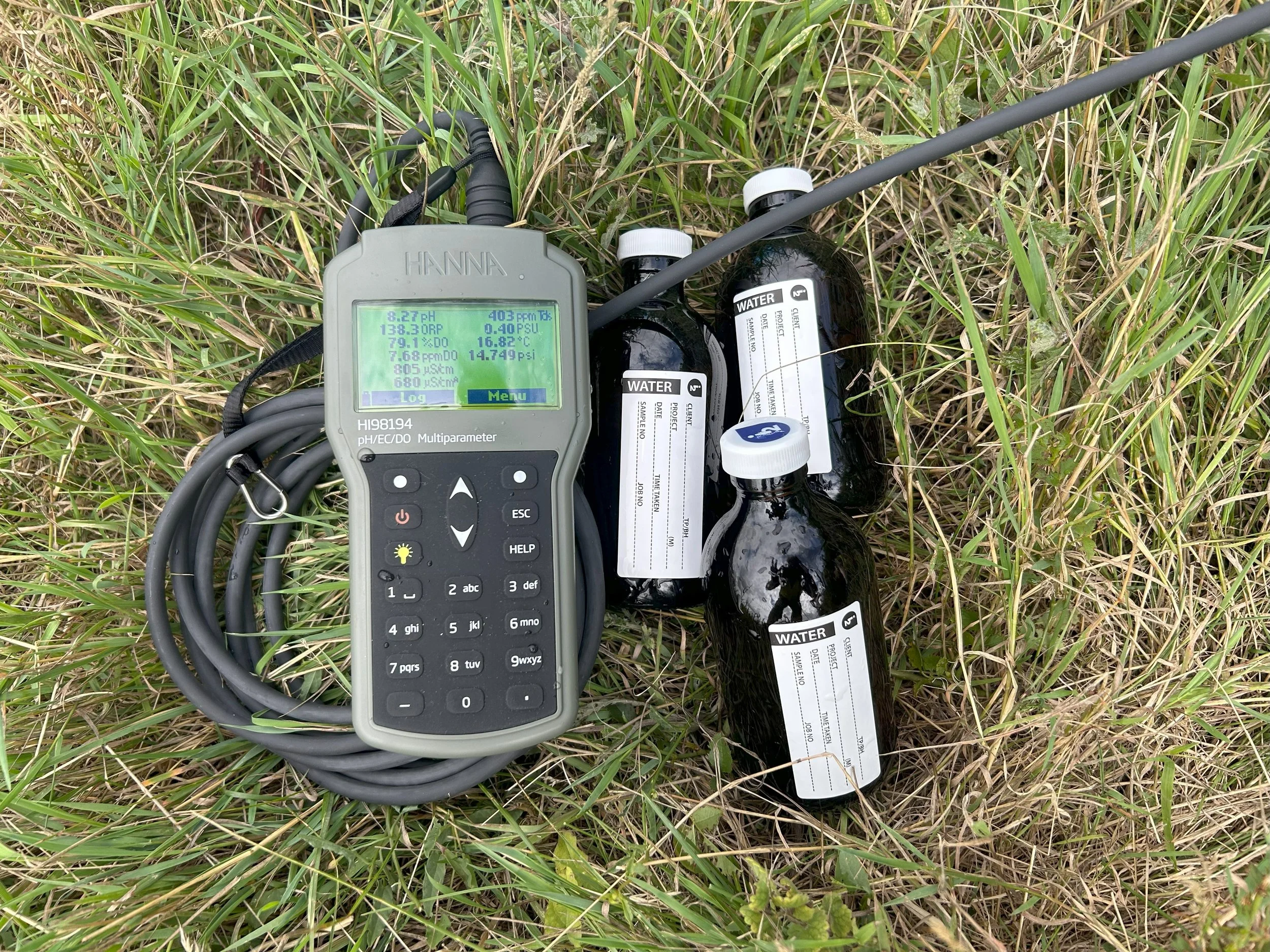 Water sampling equipment on grass, including a pH/EC/DO meter and four bottles for water testing.