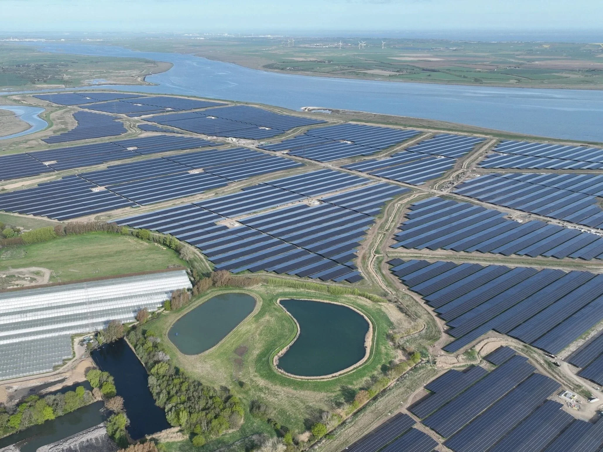 An aerial view of a large solar farm with multiple rows of solar panels, a river running in the background, and two small lakes with green surroundings in the foreground.