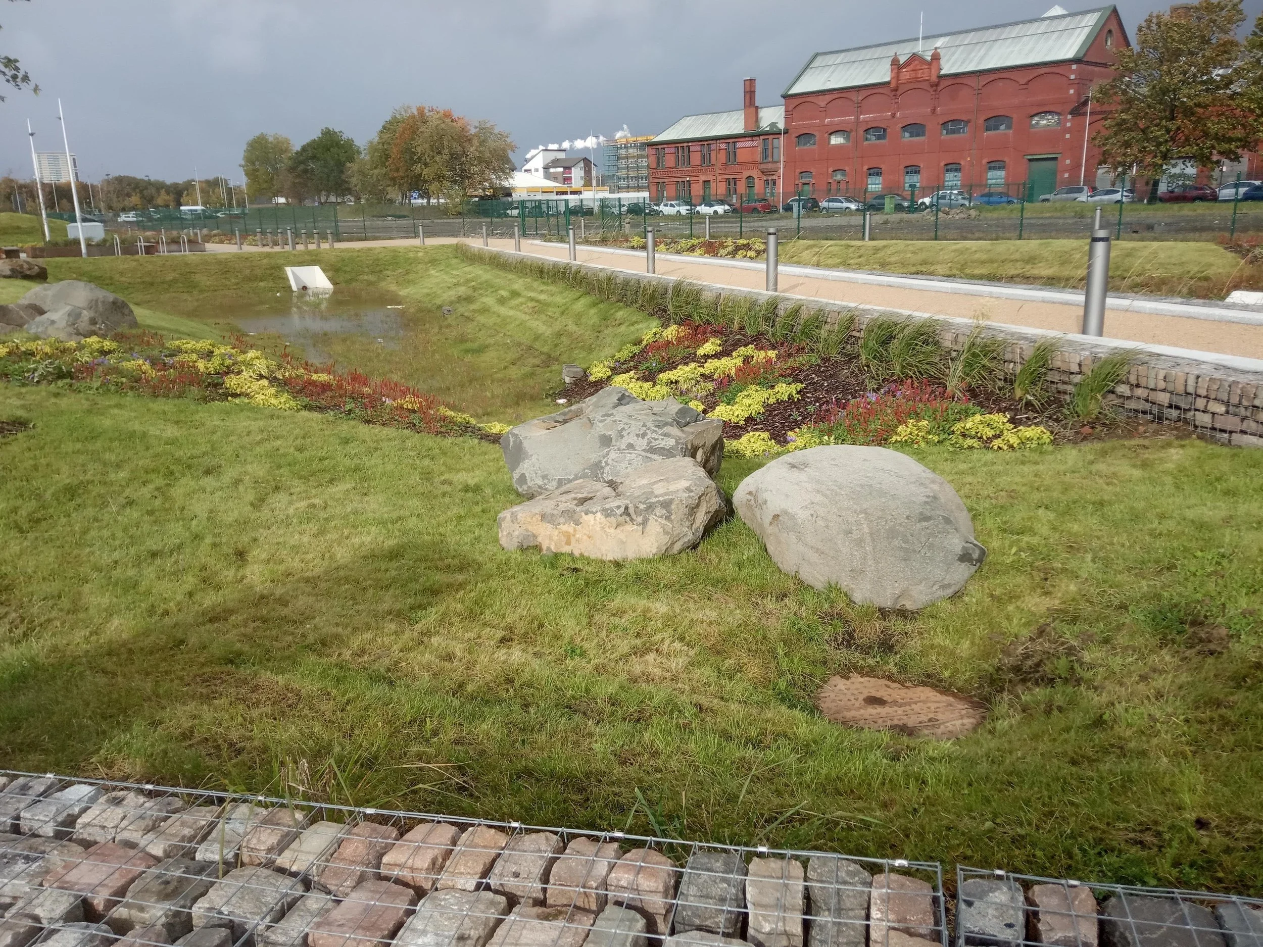 A landscaped urban park with rocks, grass, flowering plants, a small pond, a walking path, and a brick building in the background.