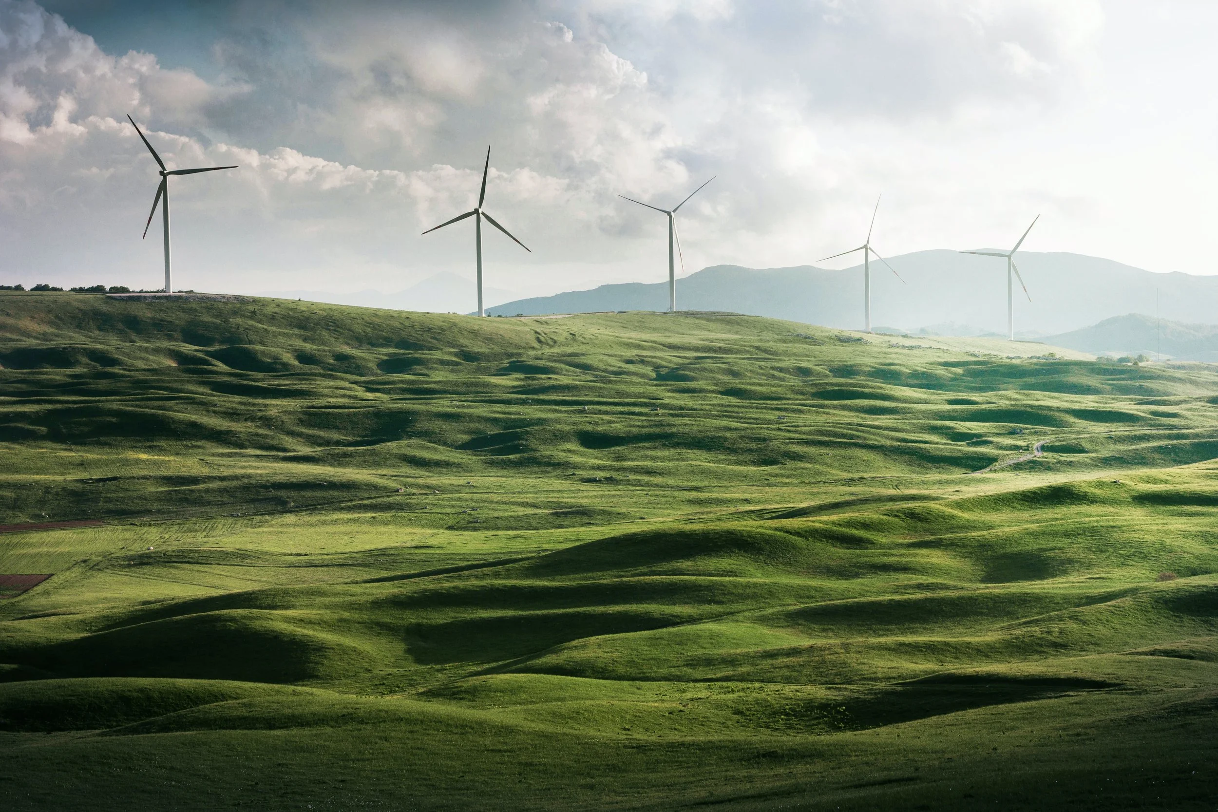 Green rolling hills with wind turbines on top, cloudy sky, distant mountains.