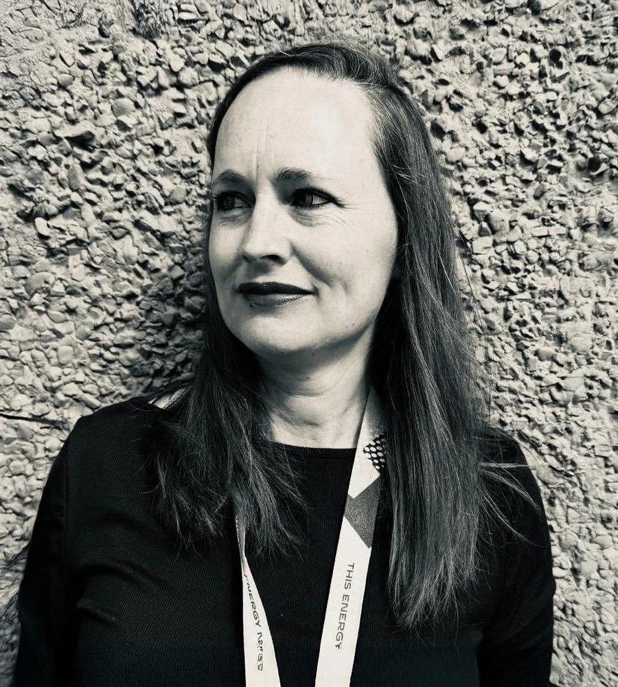 Black and white portrait of a woman with long dark hair, wearing a black top and a lanyard, standing against a textured wall.