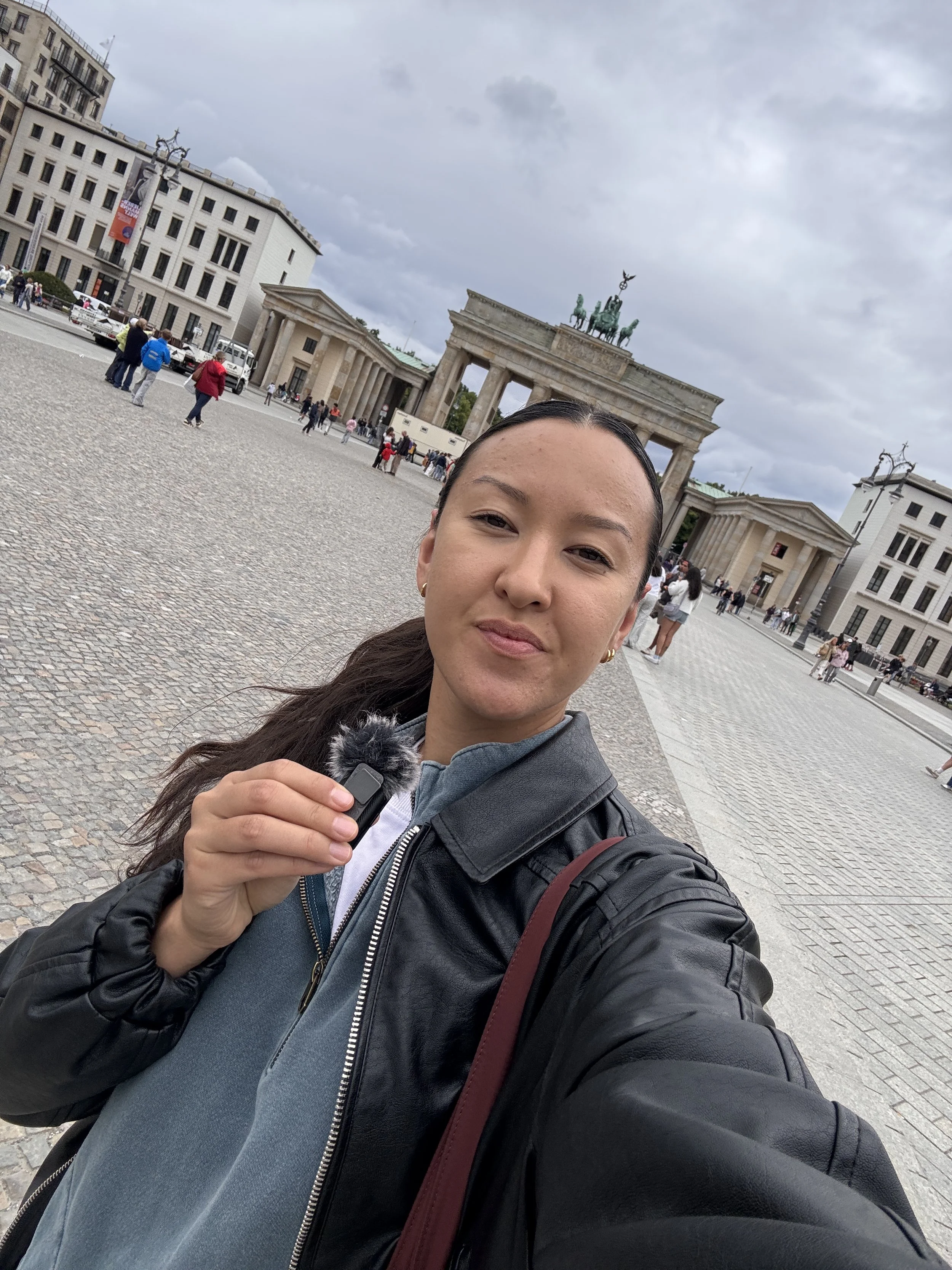 A woman taking a selfie in front of the Brandenburg Gate in Berlin, Germany, with a cloudy sky overhead.