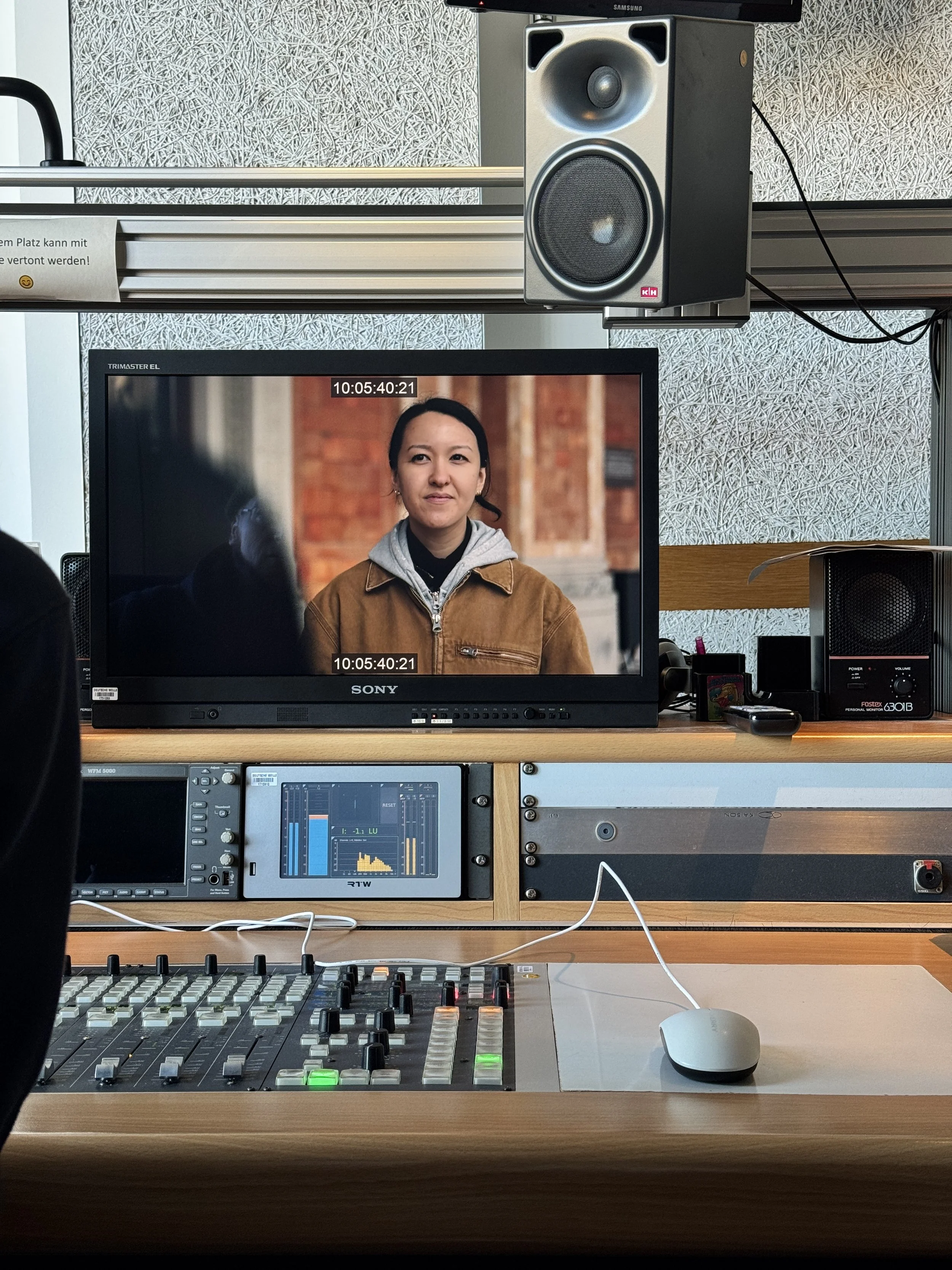 A computer workstation with an audio speaker mounted above a monitor, a Sony monitor displaying a woman, a mixing console, a keyboard, a mouse, and an external speaker in the background.