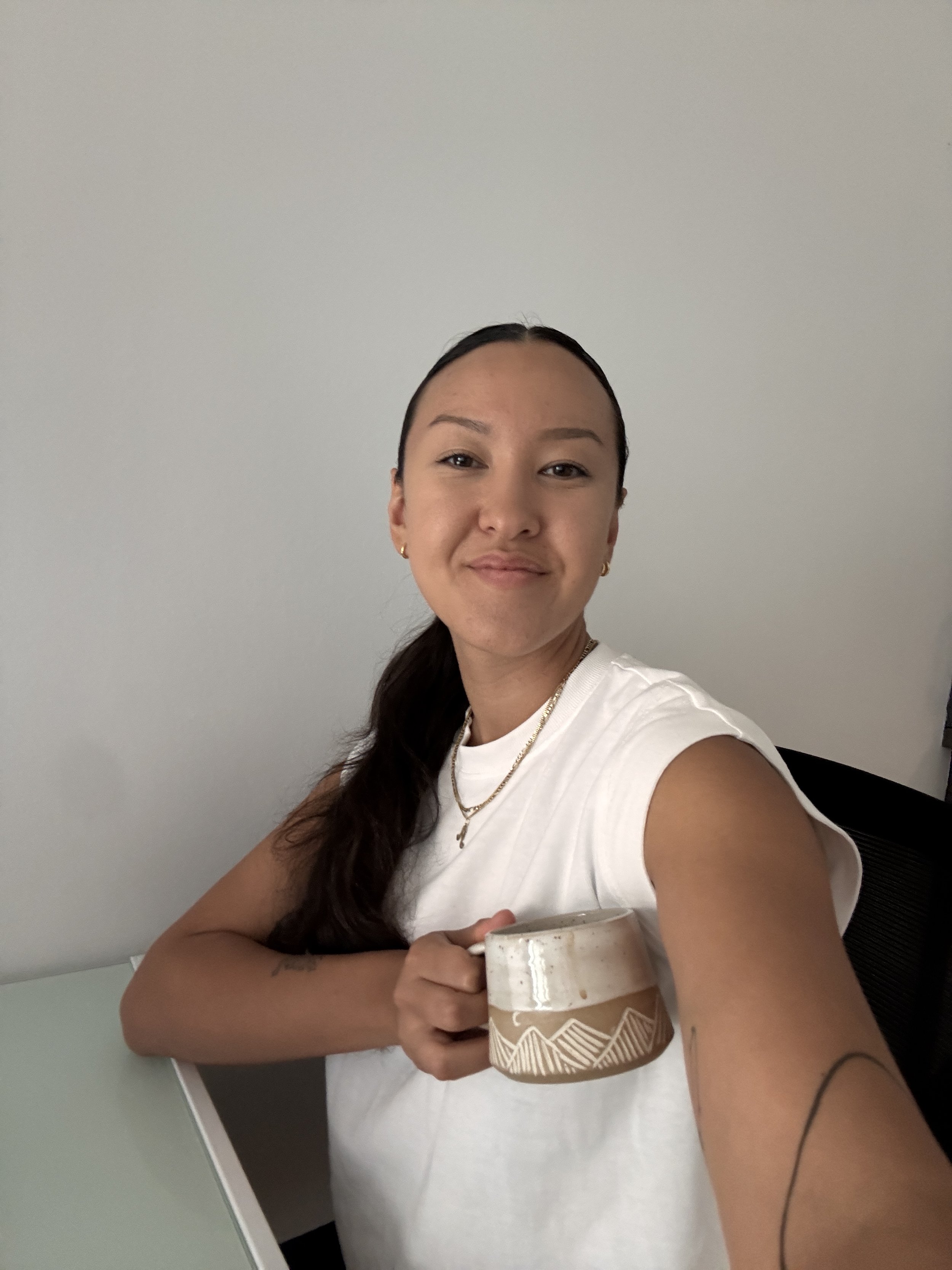 A young woman with dark, wavy hair tied back, wearing a white sleeveless top and gold jewelry, holding a ceramic mug with a geometric pattern, sitting at a table against a plain light-colored wall.