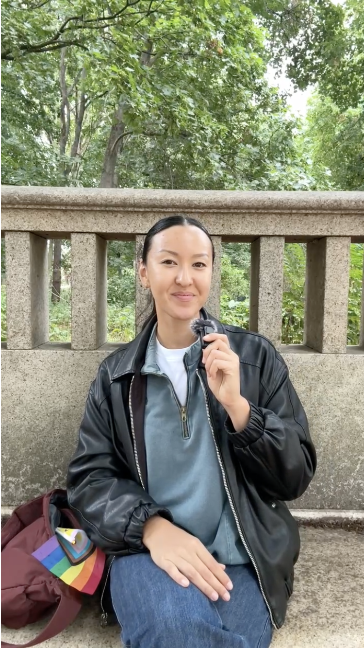 A woman sitting outdoors in front of a concrete railing, holding a small fluffy gray animal, with trees and green foliage in the background.