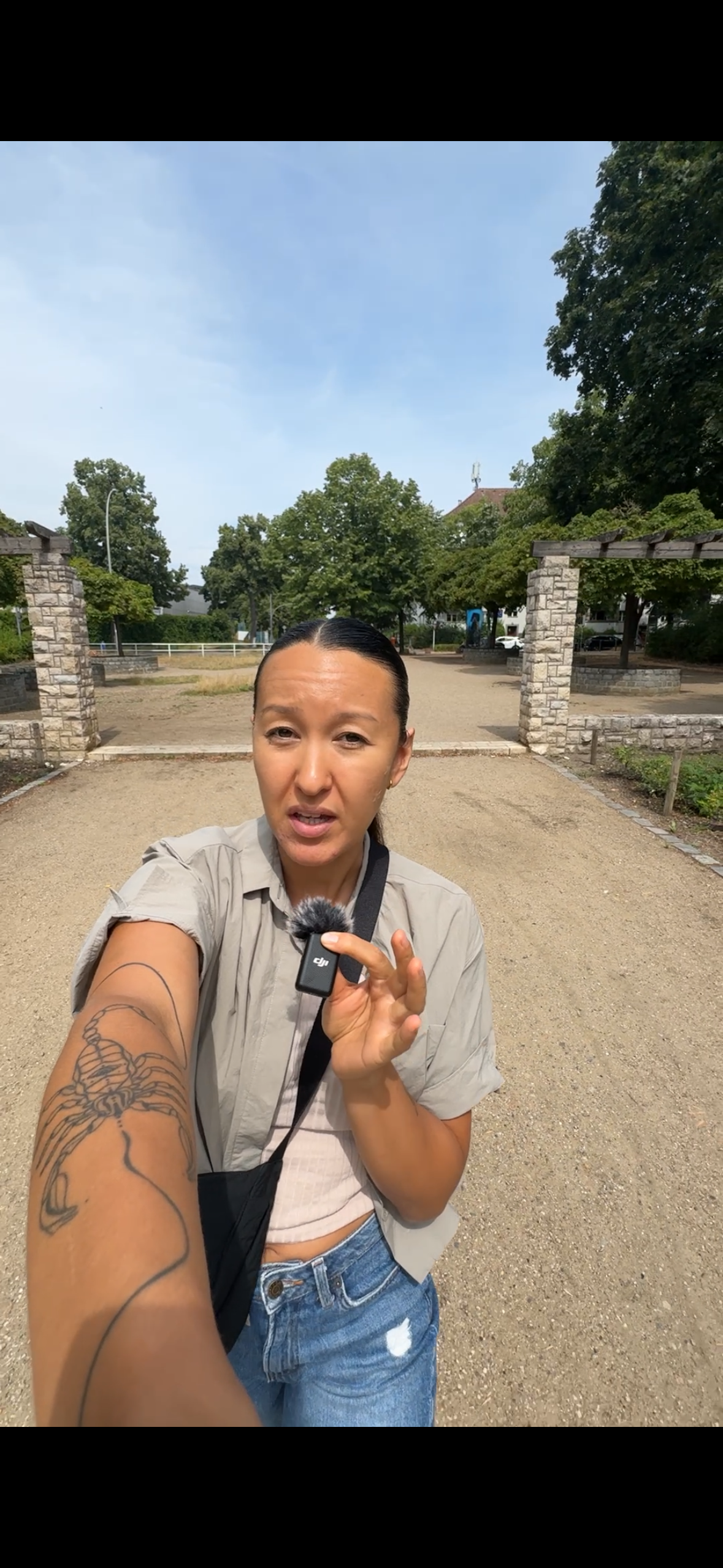 Woman taking a selfie in a park, holding a microphone, with trees and a stone archway in the background on a sunny day.