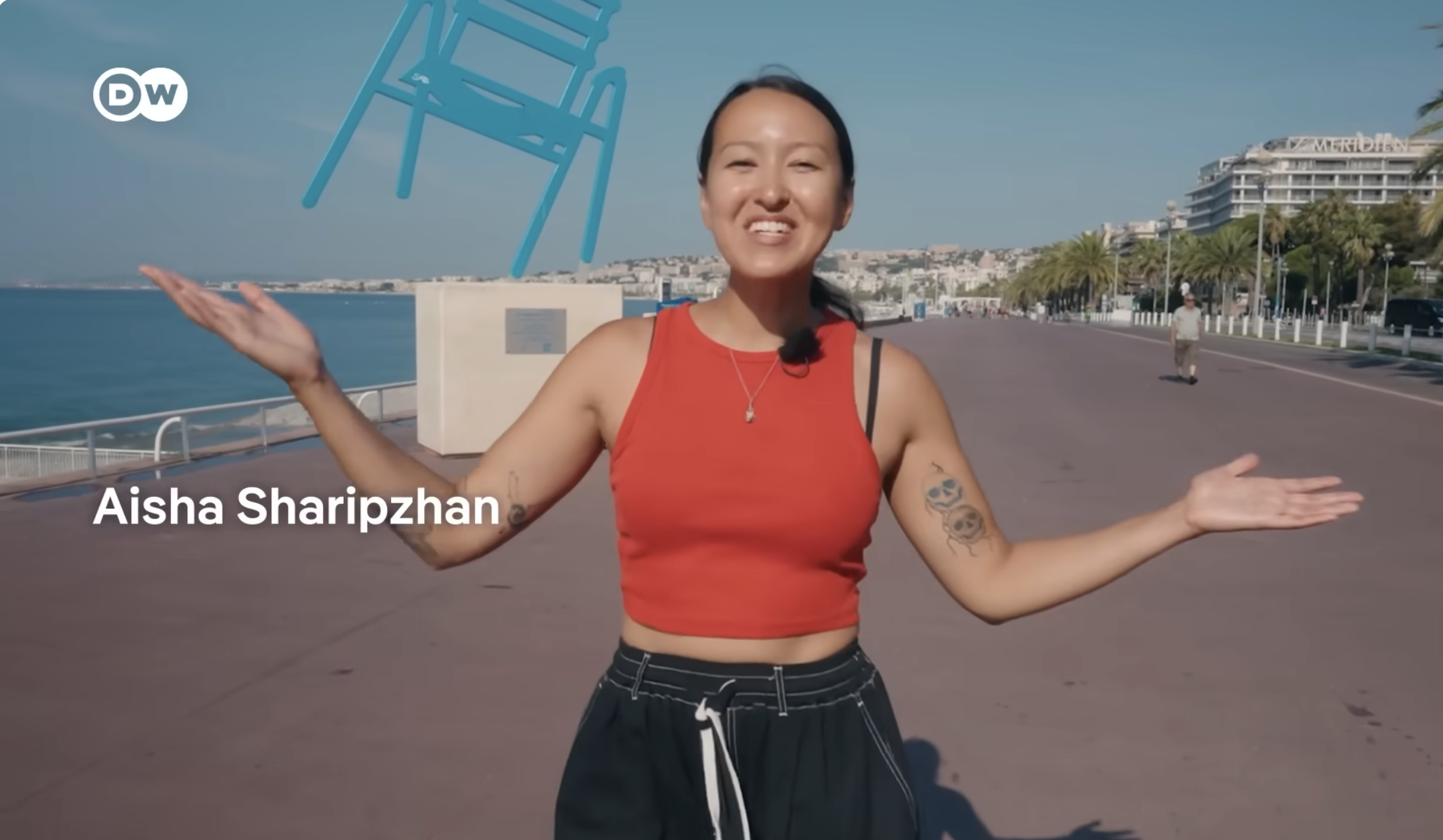 A woman in a red sleeveless top and black shorts standing outdoors near a waterfront, smiling with her arms outstretched, with a blue sky and buildings in the background.