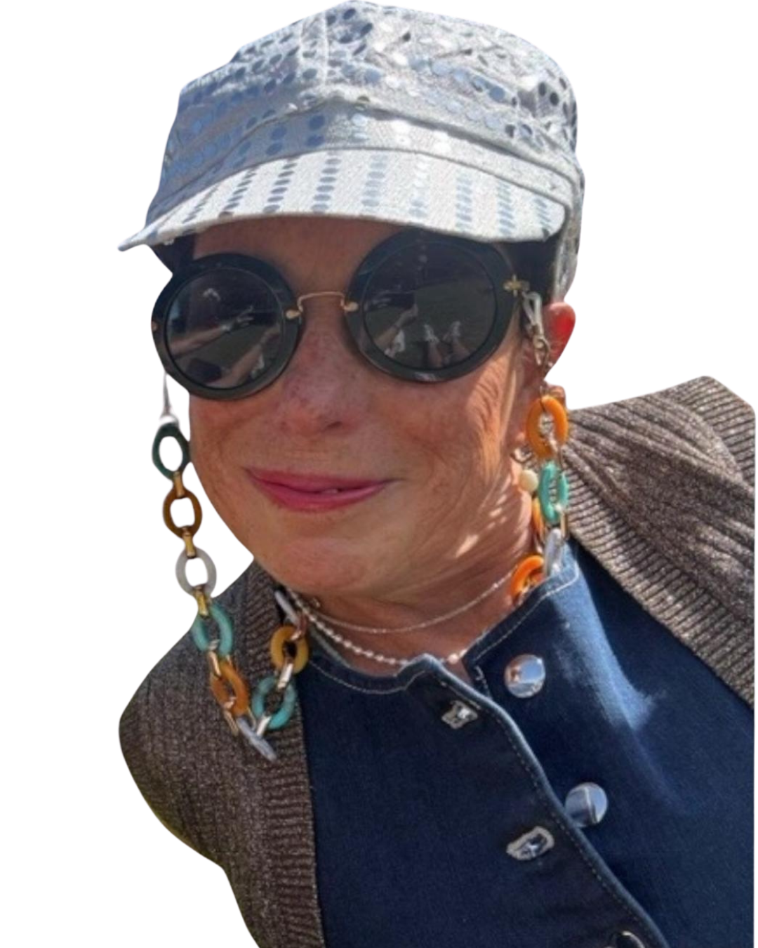 Julie Brooker is smiling, wearing dark round sunglasses, a gray and white patterned cap, colorful chain earrings, a multi-colored chain necklace, and a gray jacket over a dark blue shirt.