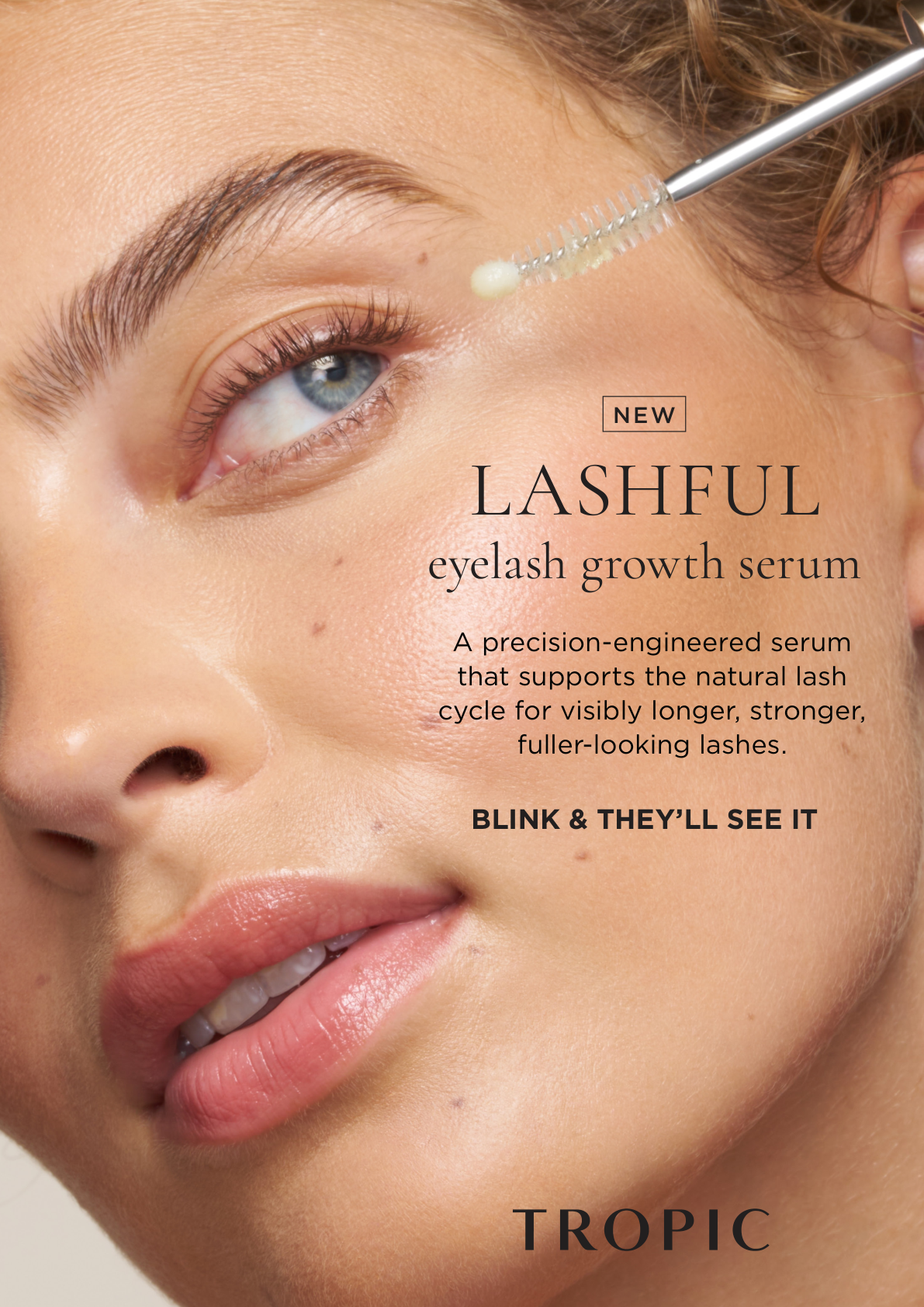 Close-up of a woman's face with blue eyes and light skin, applying eyelash growth serum with a brush near her eye.