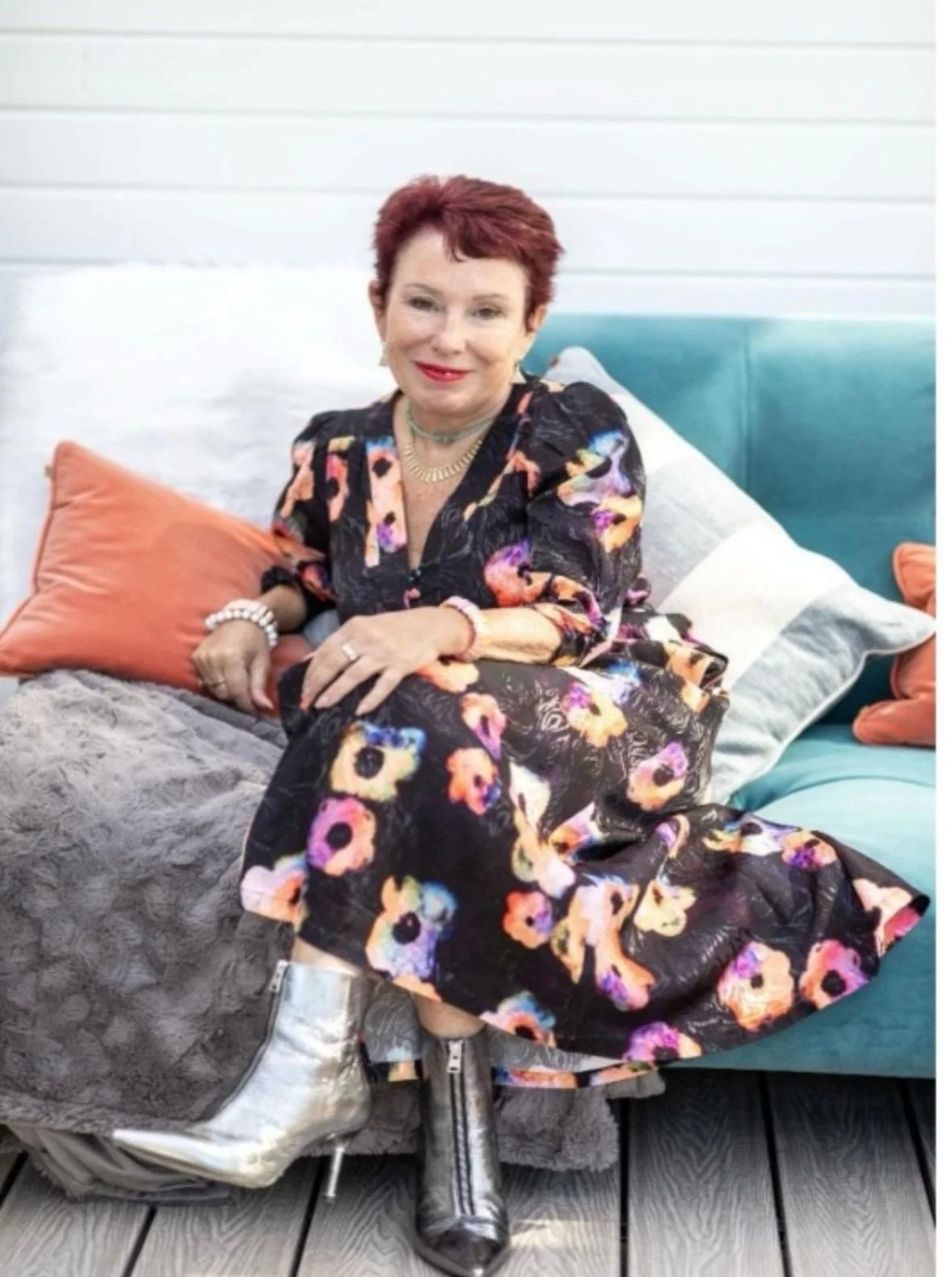 Julie Brooker wearing a floral dress, sitting on a cozy outdoor sofa with pillows, smiling at the camera.