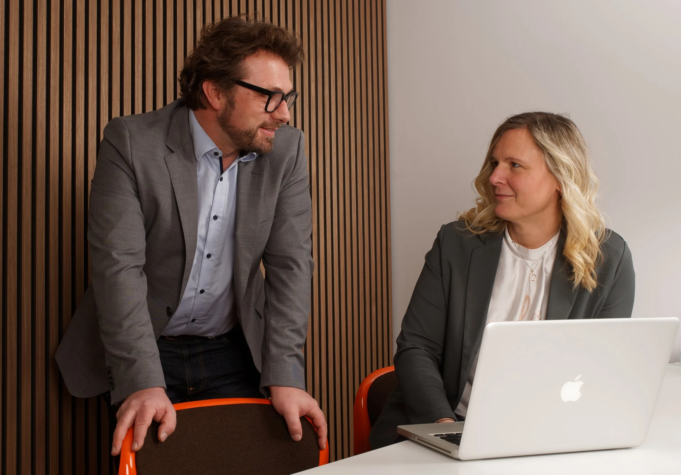 Zwei Personen, ein Mann und eine Frau, stehen und sitzen bei der Arbeit im Büro. Der Mann trägt eine graue Jacke und eine Brille, die Frau hat lange blonde Haare und trägt einen grauen Blazer. Sie schauen auf einen weißen MacBook-Laptop auf dem Tisch vor ihnen.