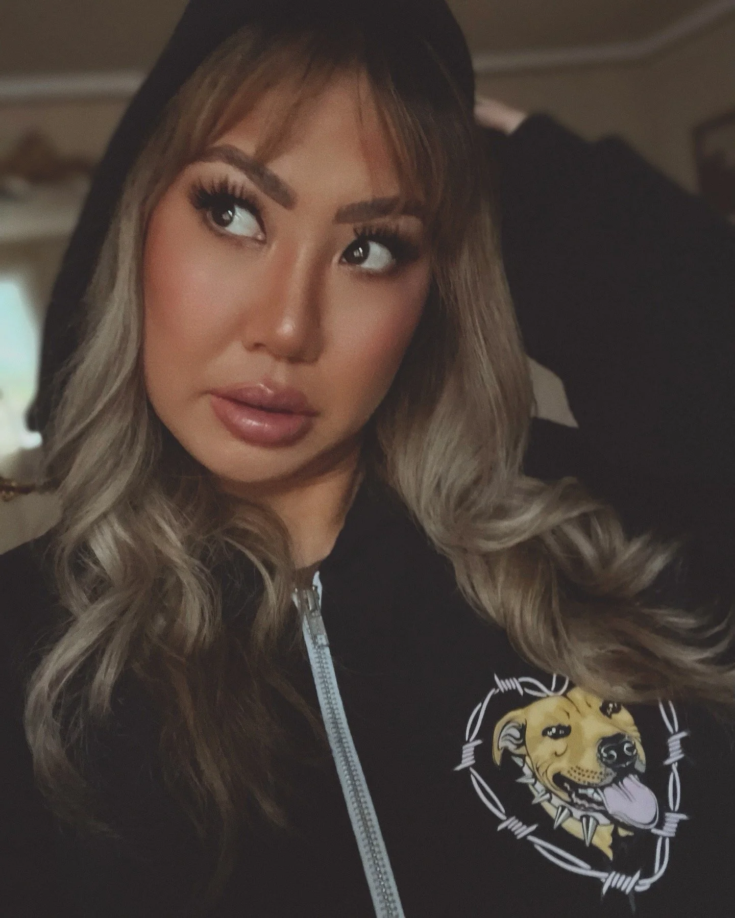 A woman with long, wavy hair and makeup in a dimly lit room. She is wearing a black hoodie with a lion graphic on it.