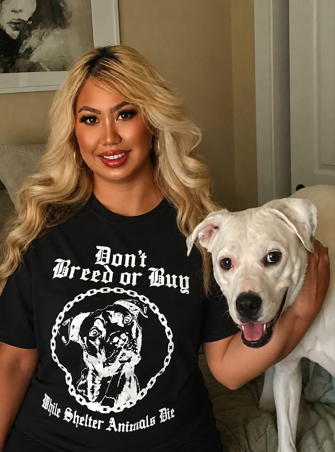A woman with long wavy blonde hair smiling, wearing a black T-shirt with a graphic of a dog and the text 'Don't Breed or Buy, White Shelter Animals Die,' holding a white dog with a black nose and dark eyes, in a cozy room.