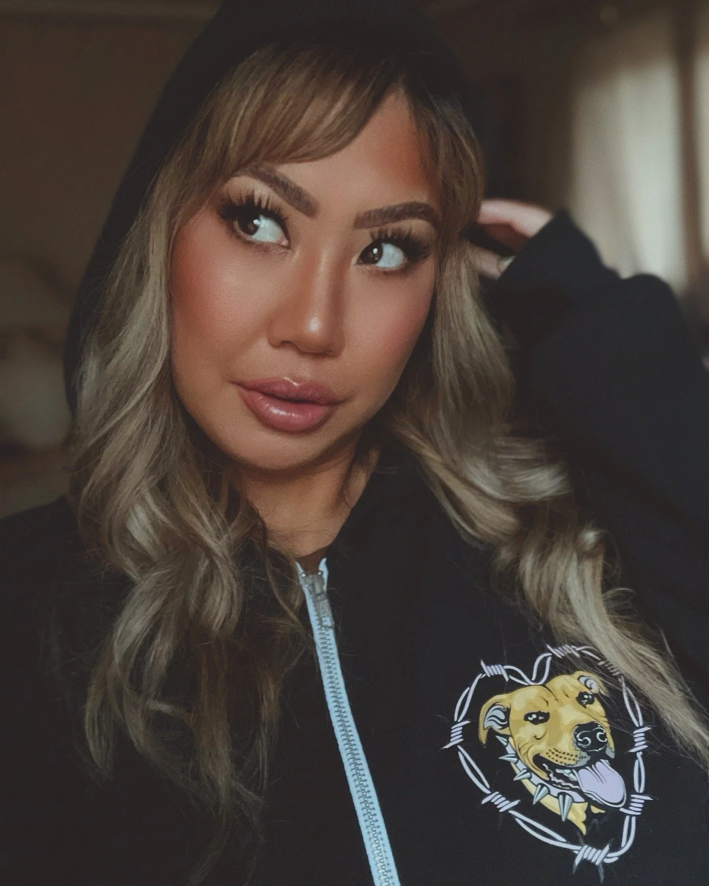A woman with long wavy blonde hair, wearing makeup with thick eyelashes and glossy lips, dressed in a black zip-up hoodie with a lion graphic on the chest. She is touching her head with her right hand, and the background is blurred.